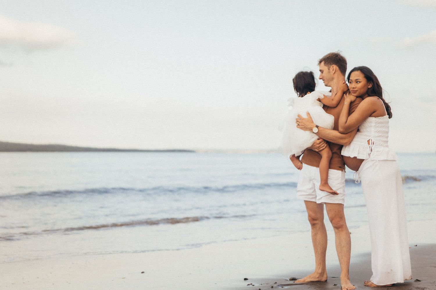 A happy family of three enjoys a romantic beach sunset, with parents holding their toddler near the ocean shore.