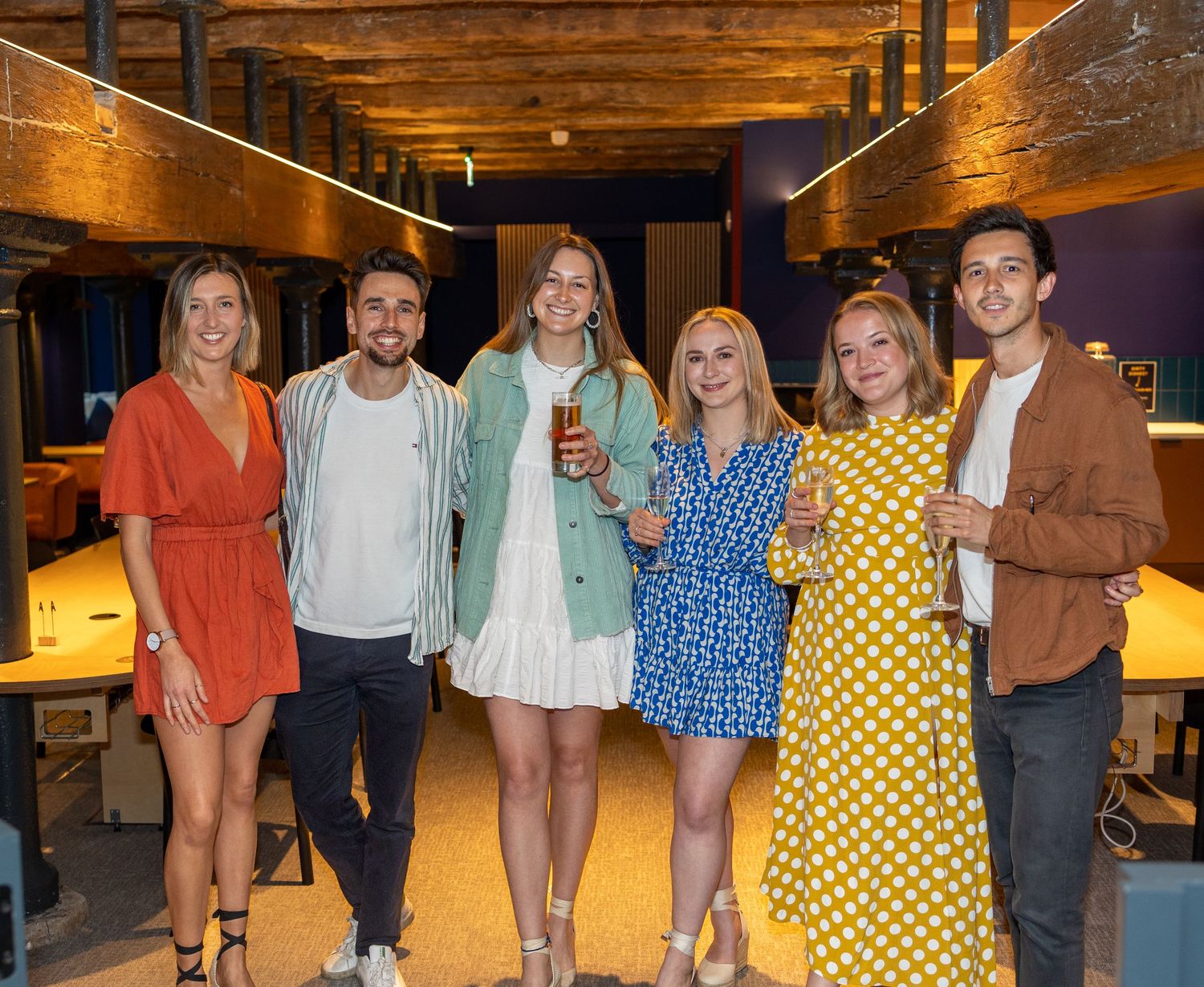 Six young adults pose together smiling at an indoor party venue with warm lighting.