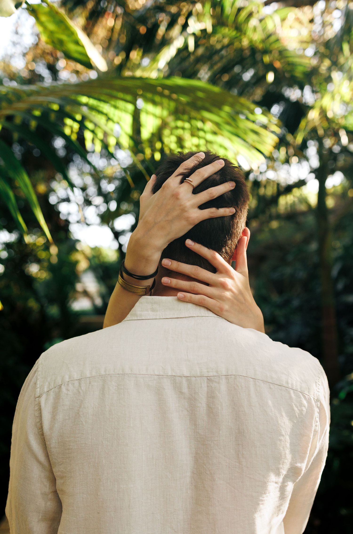 Back view of man in white linen shirt with hand resting on head, surrounded by lush tropical greenery.