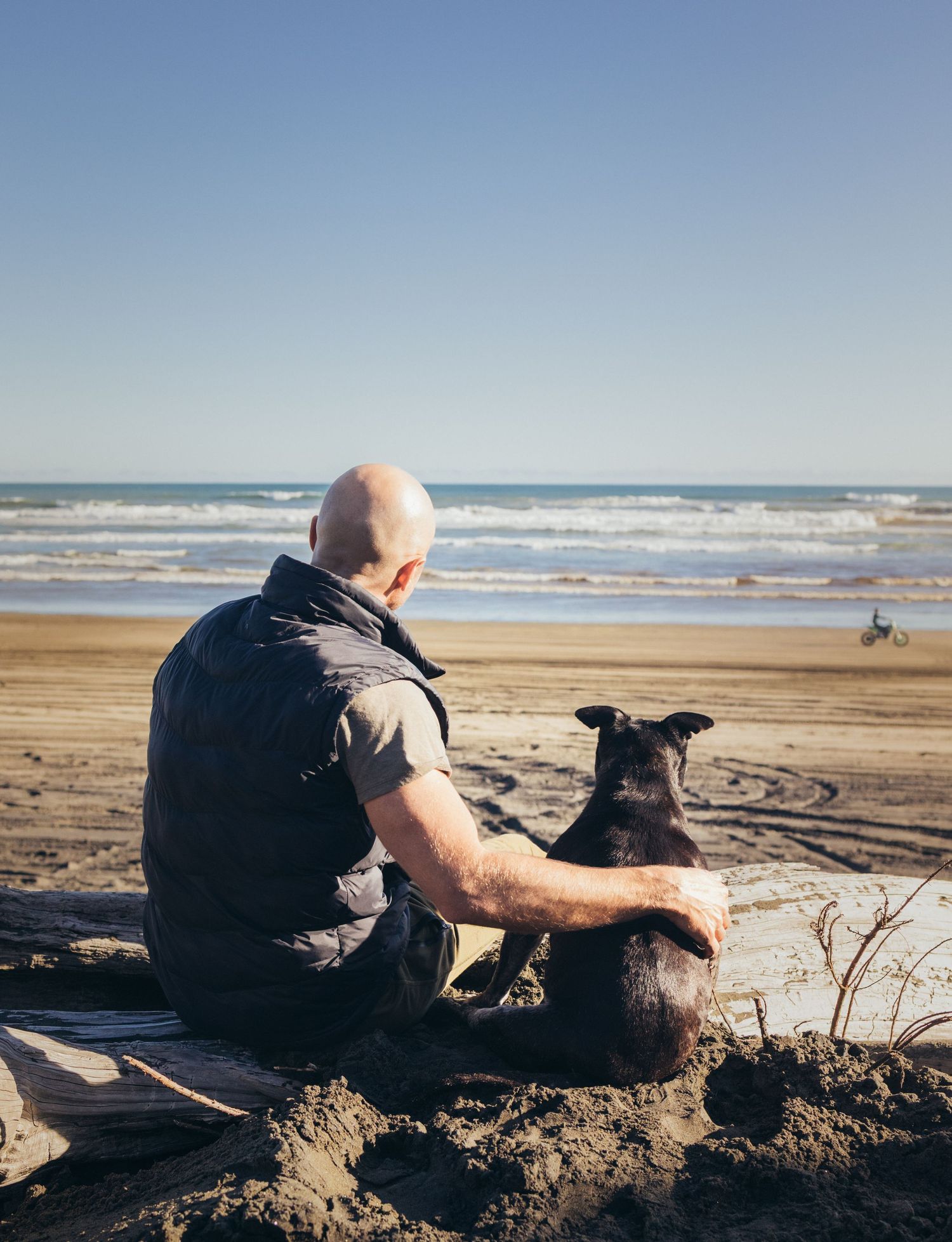 Bald man sitting on beach sand with black dog looking out at ocean waves together.