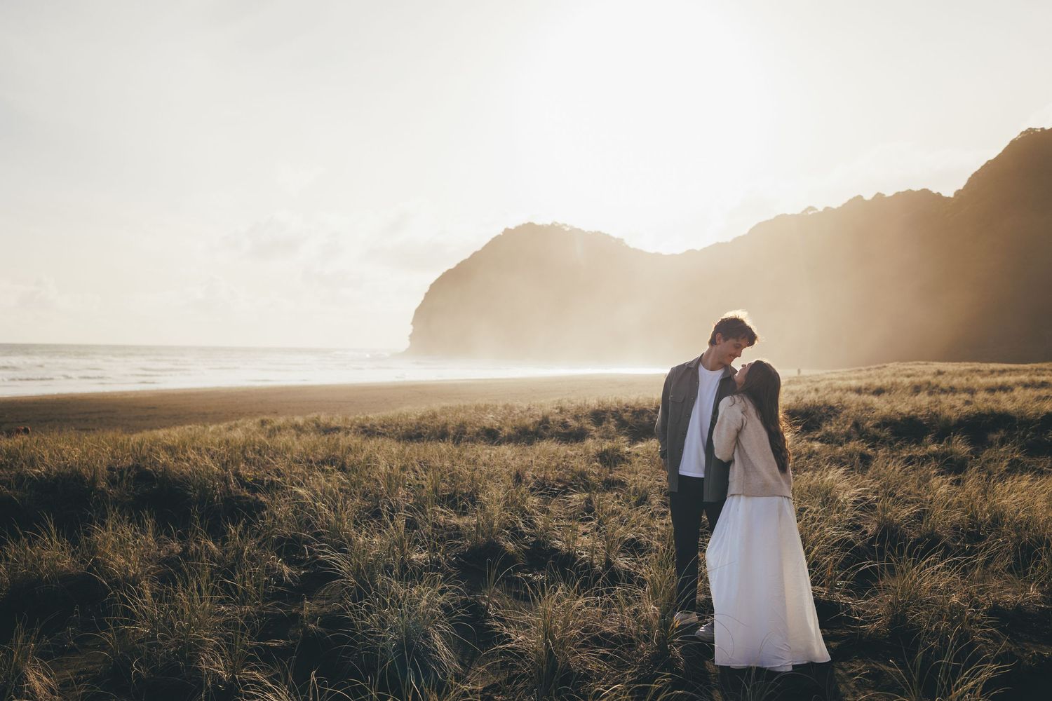 A couple shares a romantic kiss on a misty beach at golden hour, surrounded by tall grass and dramatic coastal cliffs.