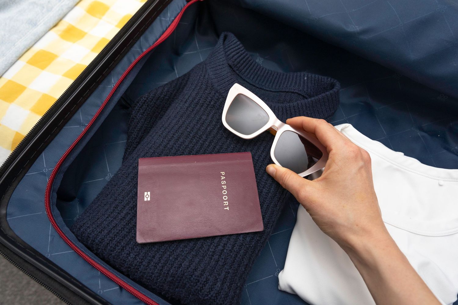 Hand placing white sunglasses next to a passport on a navy sweater inside an open travel suitcase.