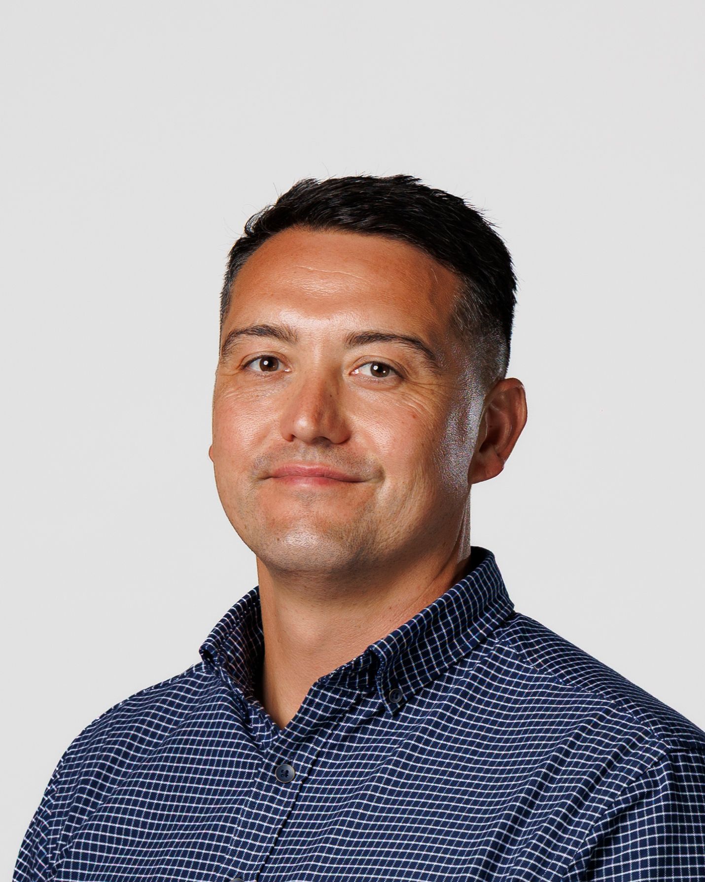 Middle-aged man with short dark hair wearing a navy checkered shirt smiling against a gray background.