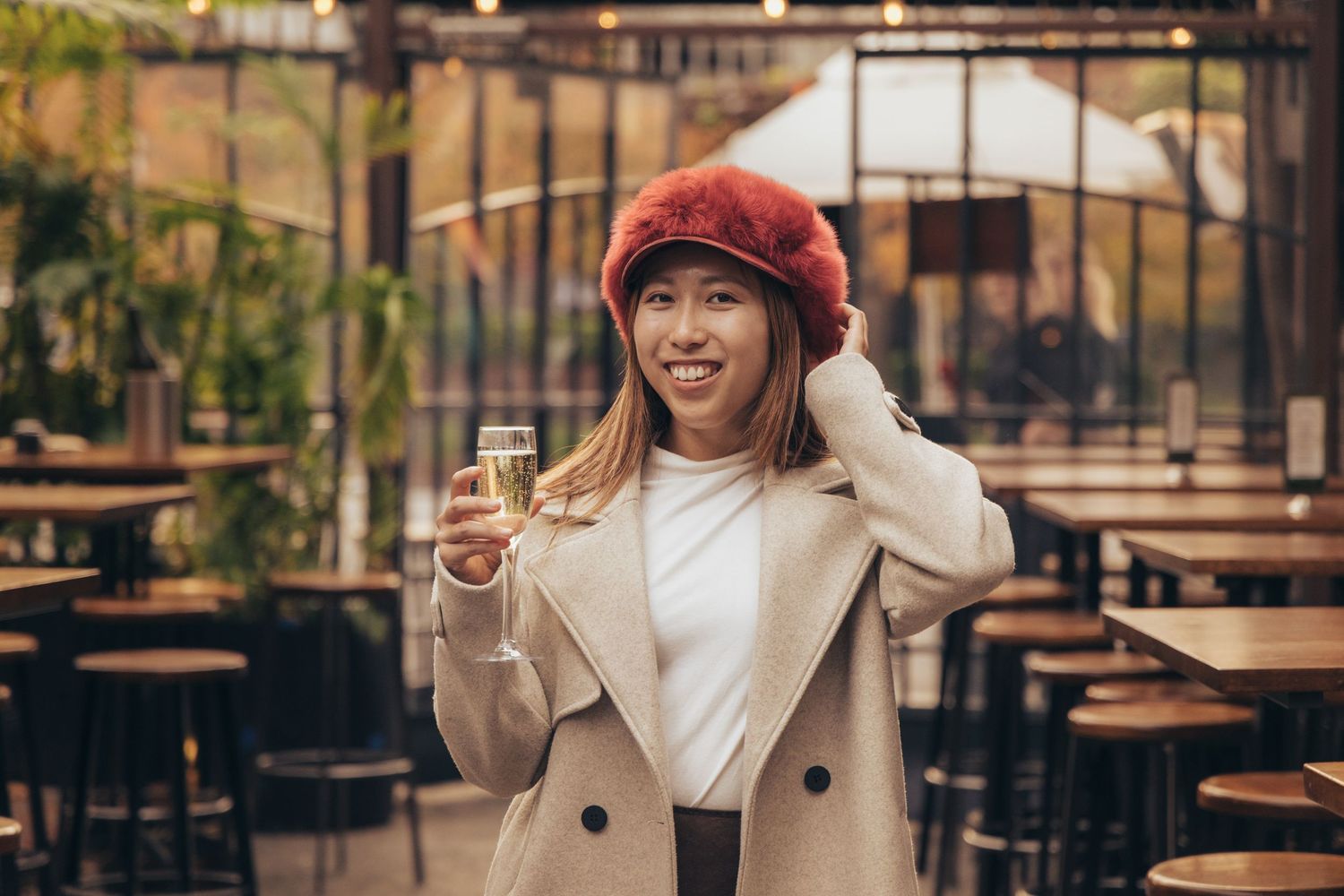 Smiling woman in red beret and beige coat holding champagne glass at outdoor cafe restaurant.