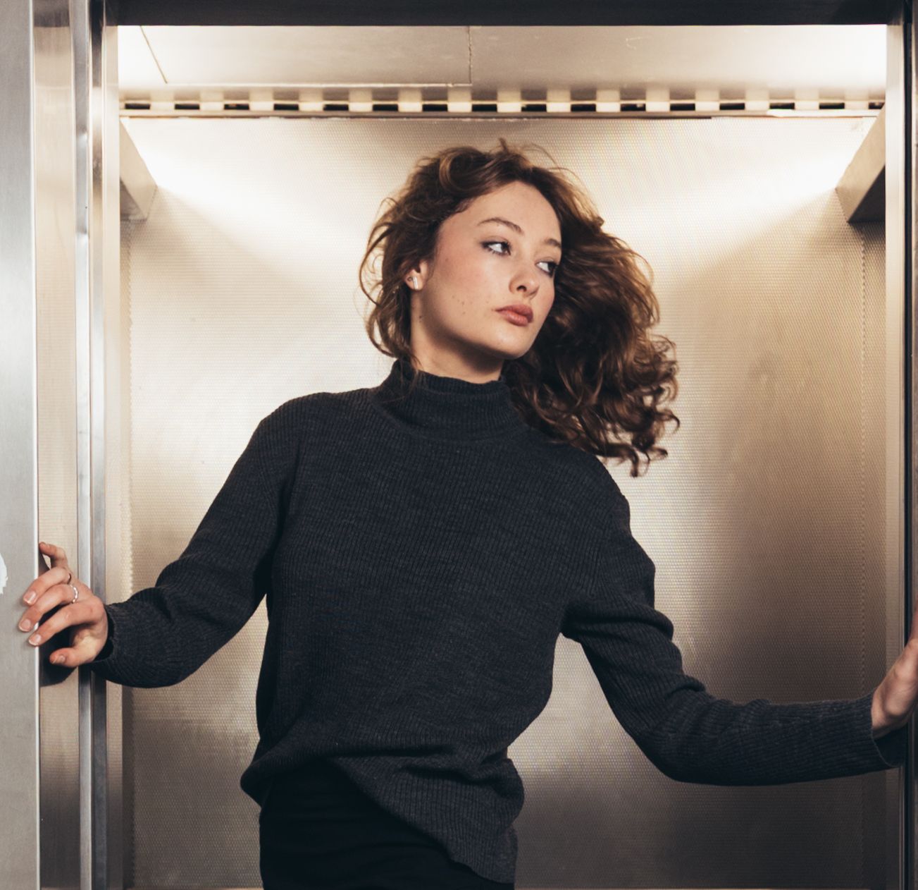 Young woman with flowing auburn hair standing between open elevator doors in warm light.