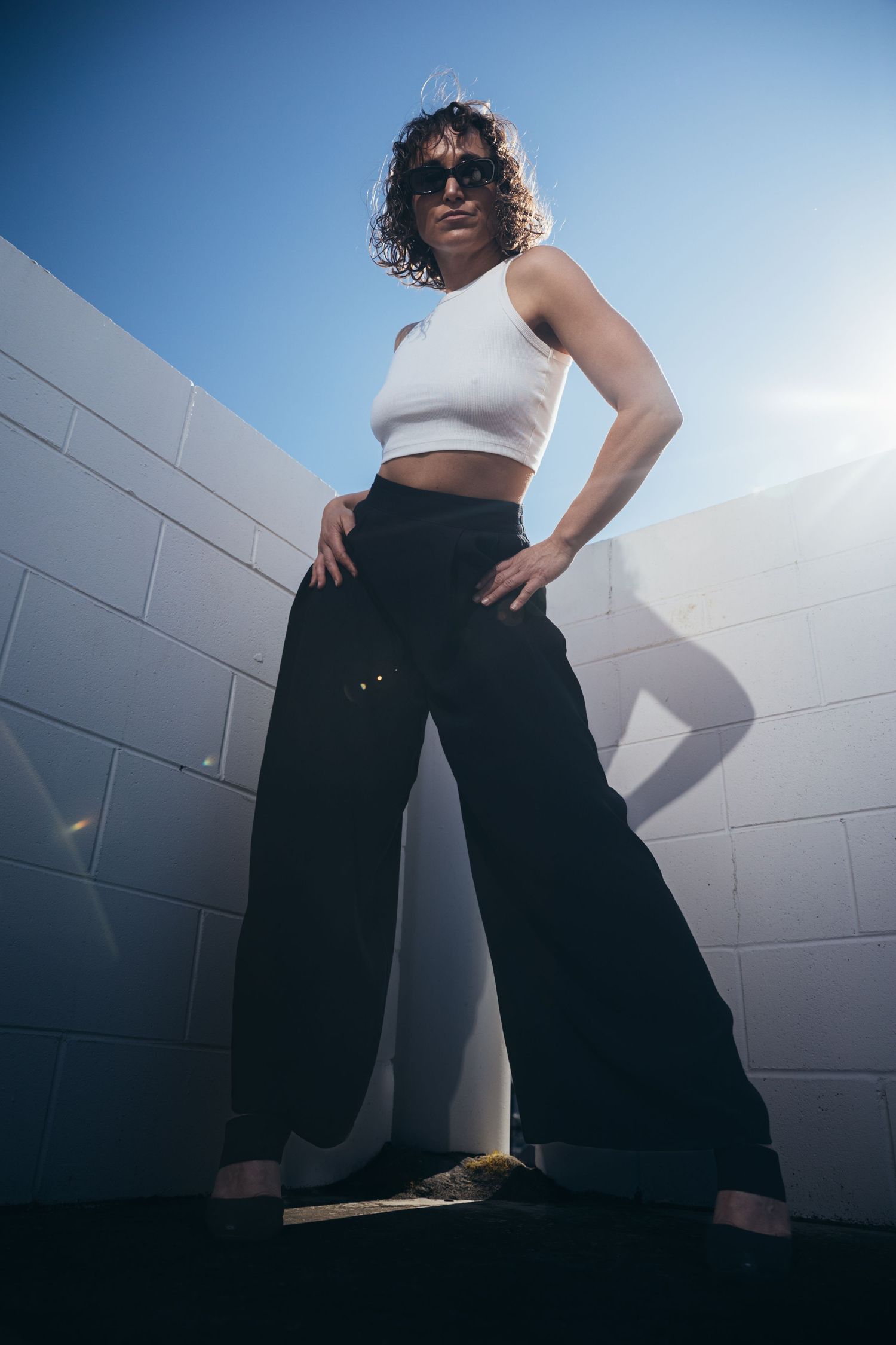 Confident woman in white crop top and black wide-leg pants poses against white brick wall in bright sunlight.