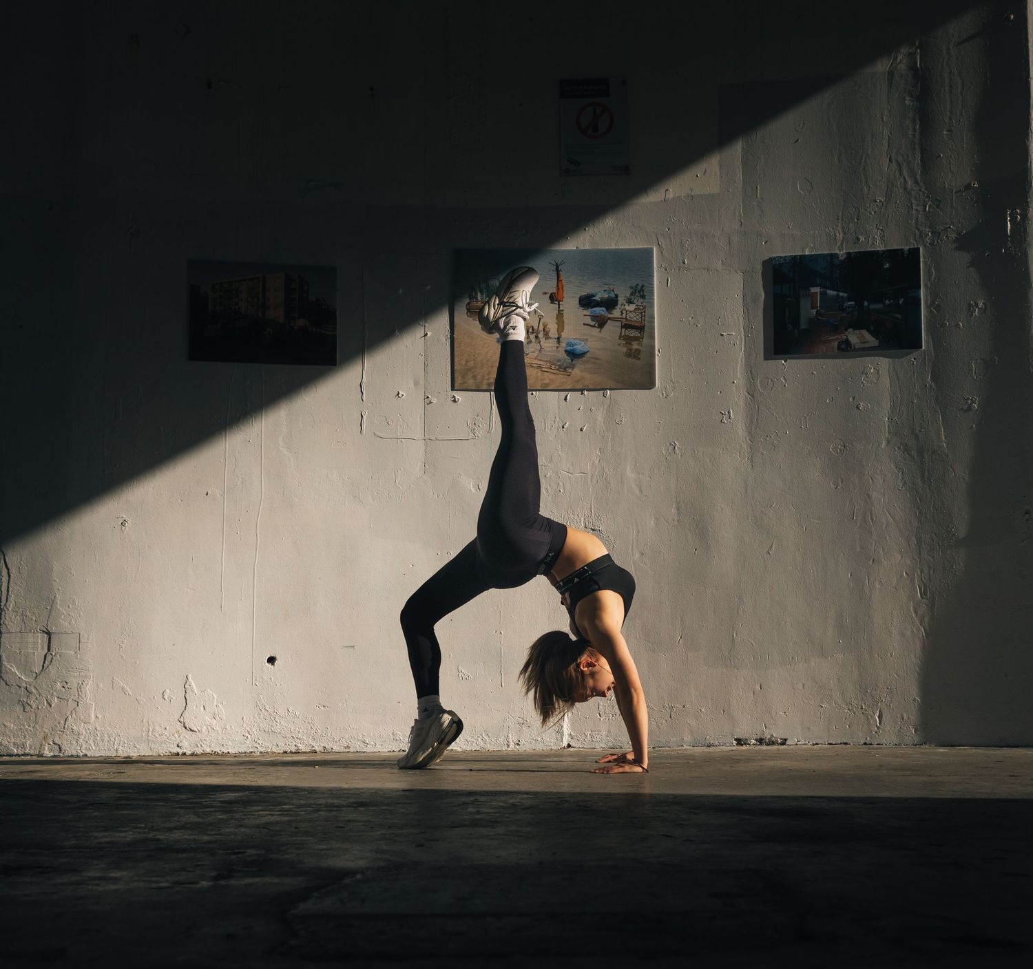 Woman in black activewear performing a one-handed handstand split against a white wall indoors.