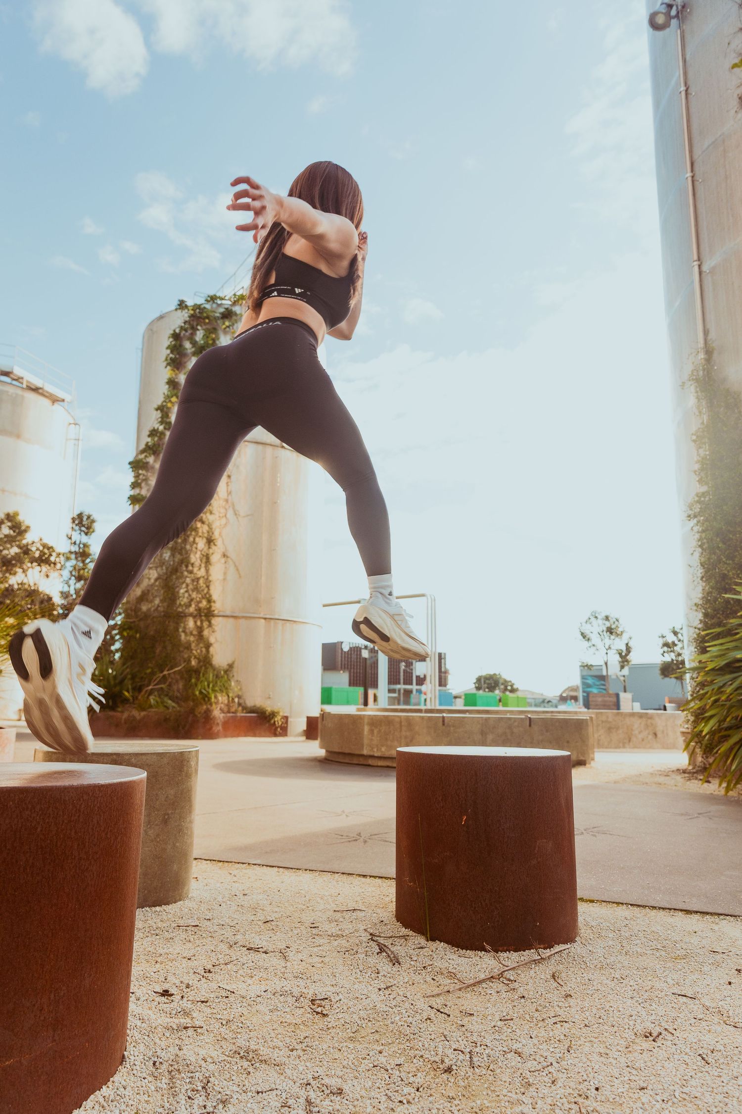 Fit woman in black activewear leaping between concrete pillars outdoors in an urban setting.