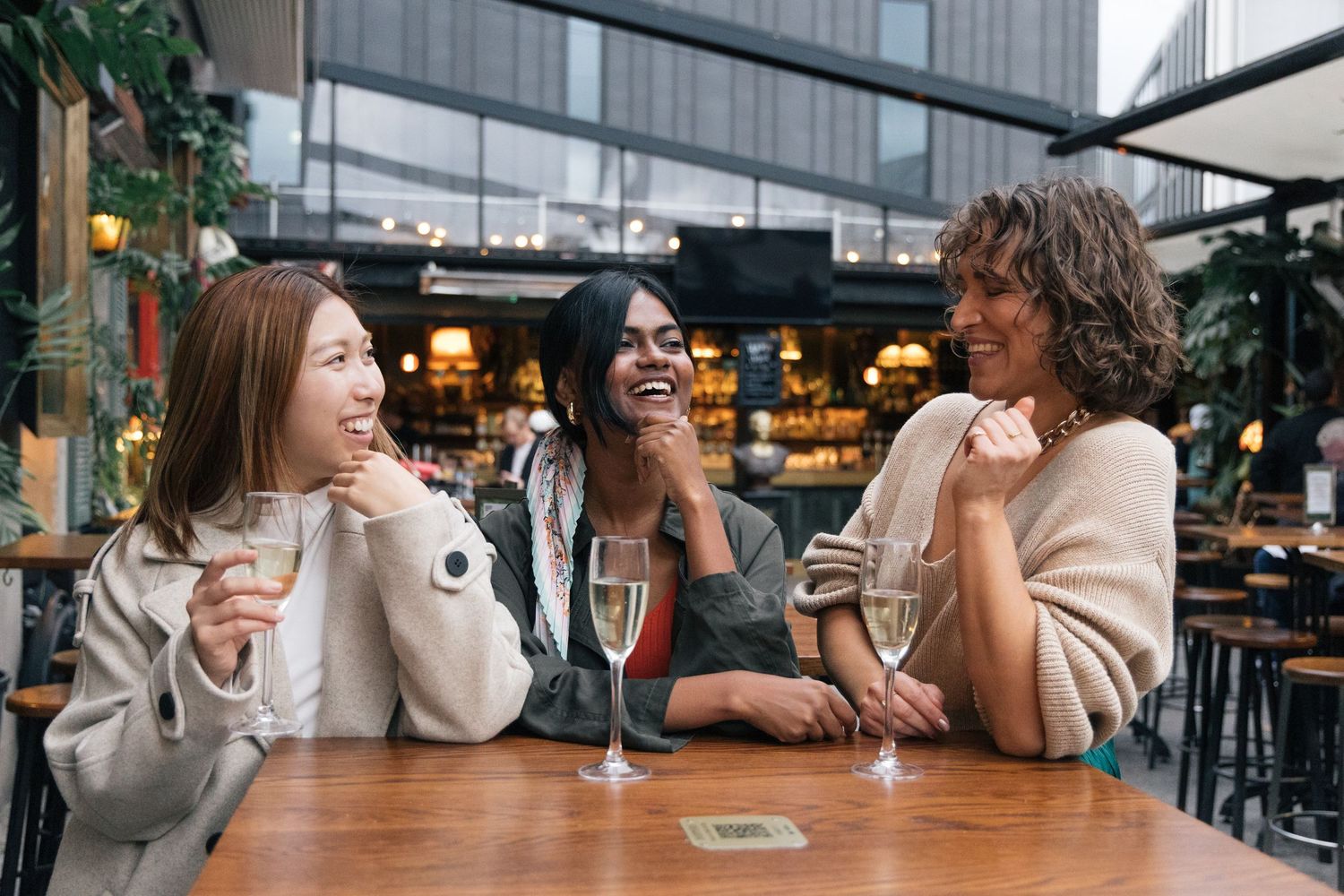 Three diverse women laughing and enjoying champagne at an outdoor cafe table together.