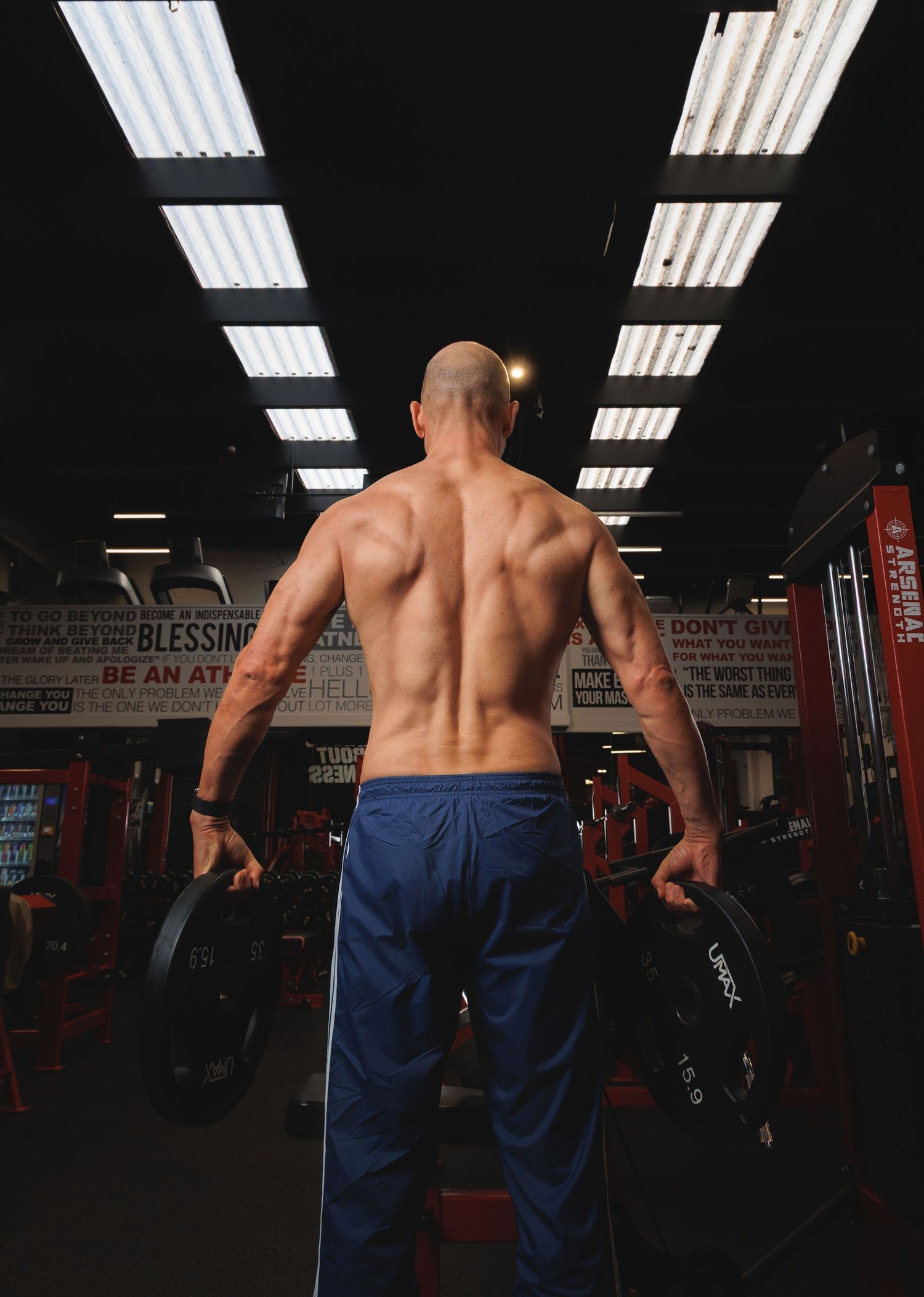 Muscular shirtless man holding weight plates in a motivational gym with red decor and bright overhead lights.