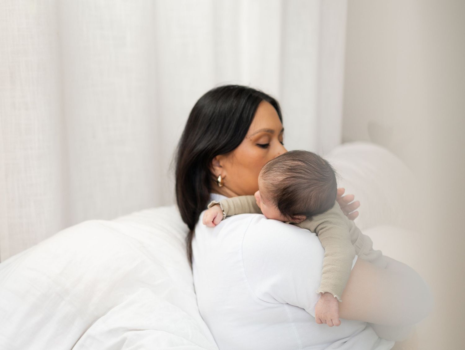 A mother with dark hair tenderly kisses her newborn baby while resting on a white bed.
