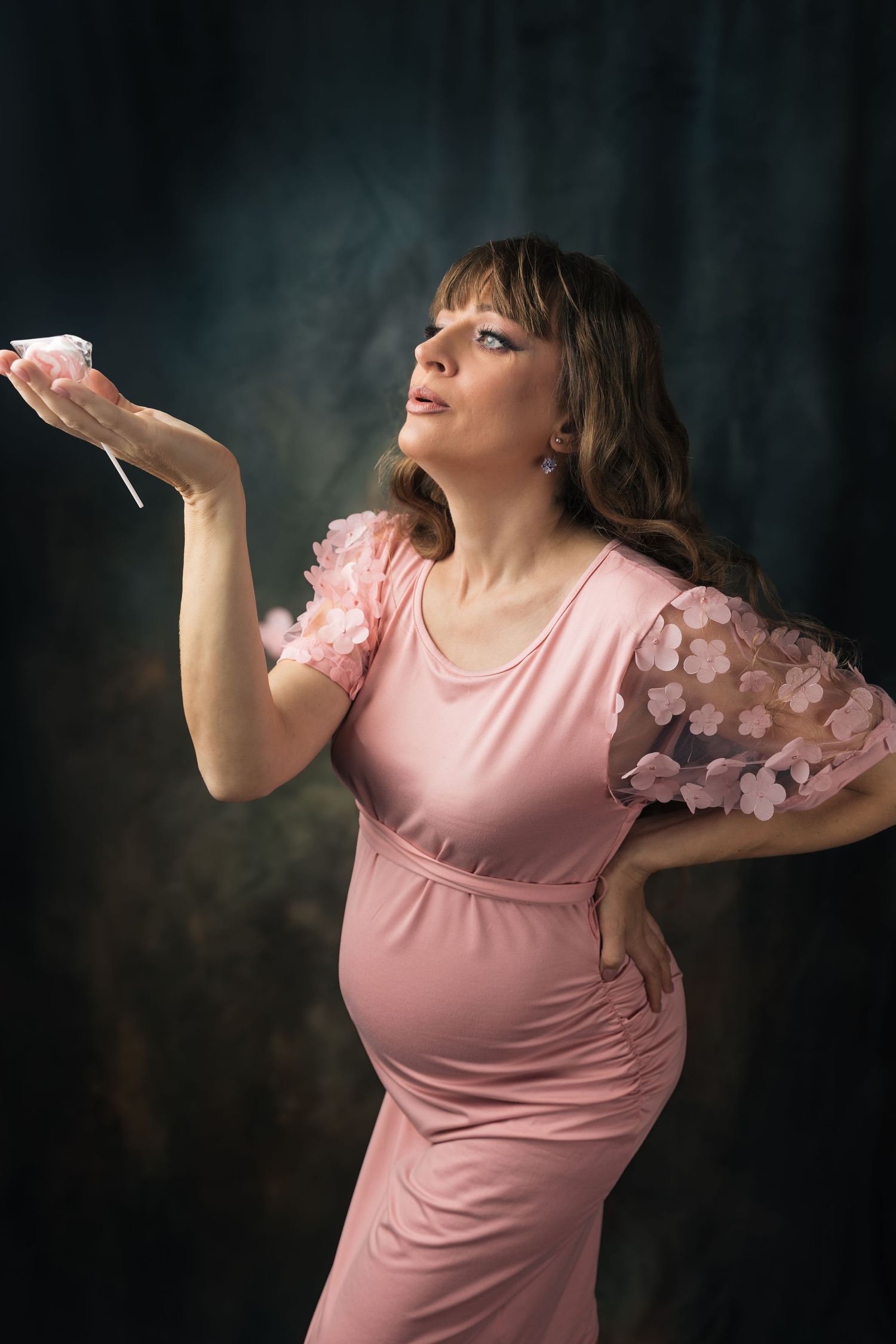 Pregnant woman in pink floral dress holding a cupcake, posing against dark backdrop.