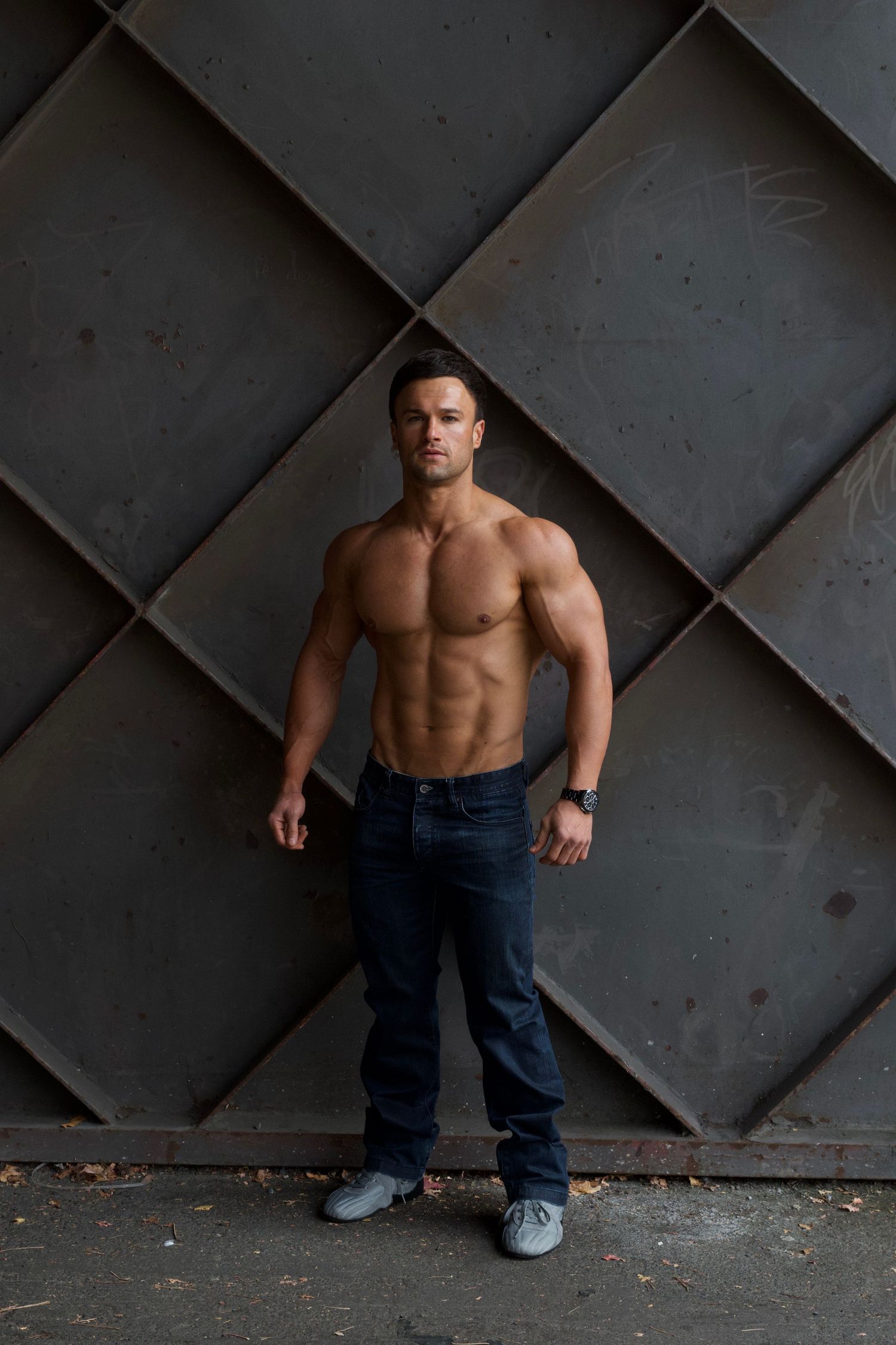 Muscular shirtless man in dark jeans standing confidently against geometric metal wall backdrop.