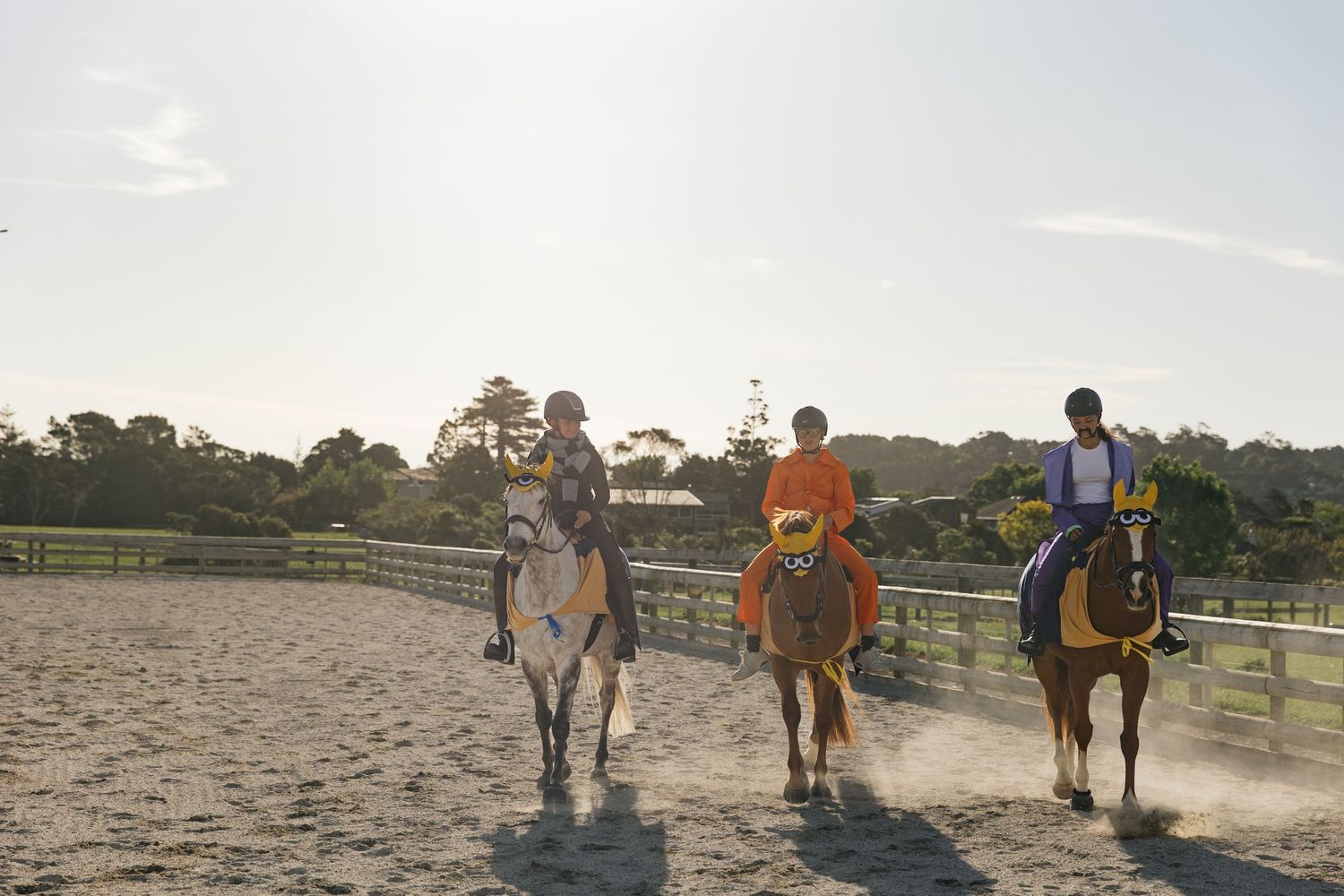 Three riders on horseback trotting through a sandy equestrian arena at golden hour.