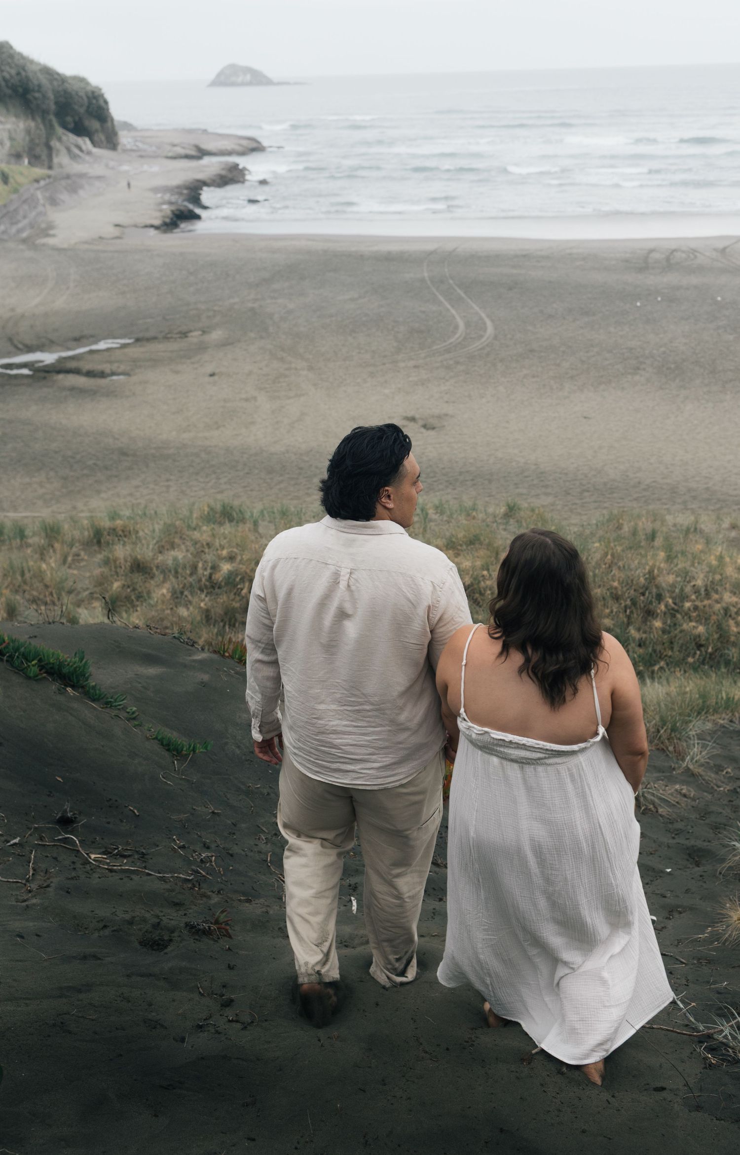 Couple walking hand in hand along a moody black sand beach with ocean views.