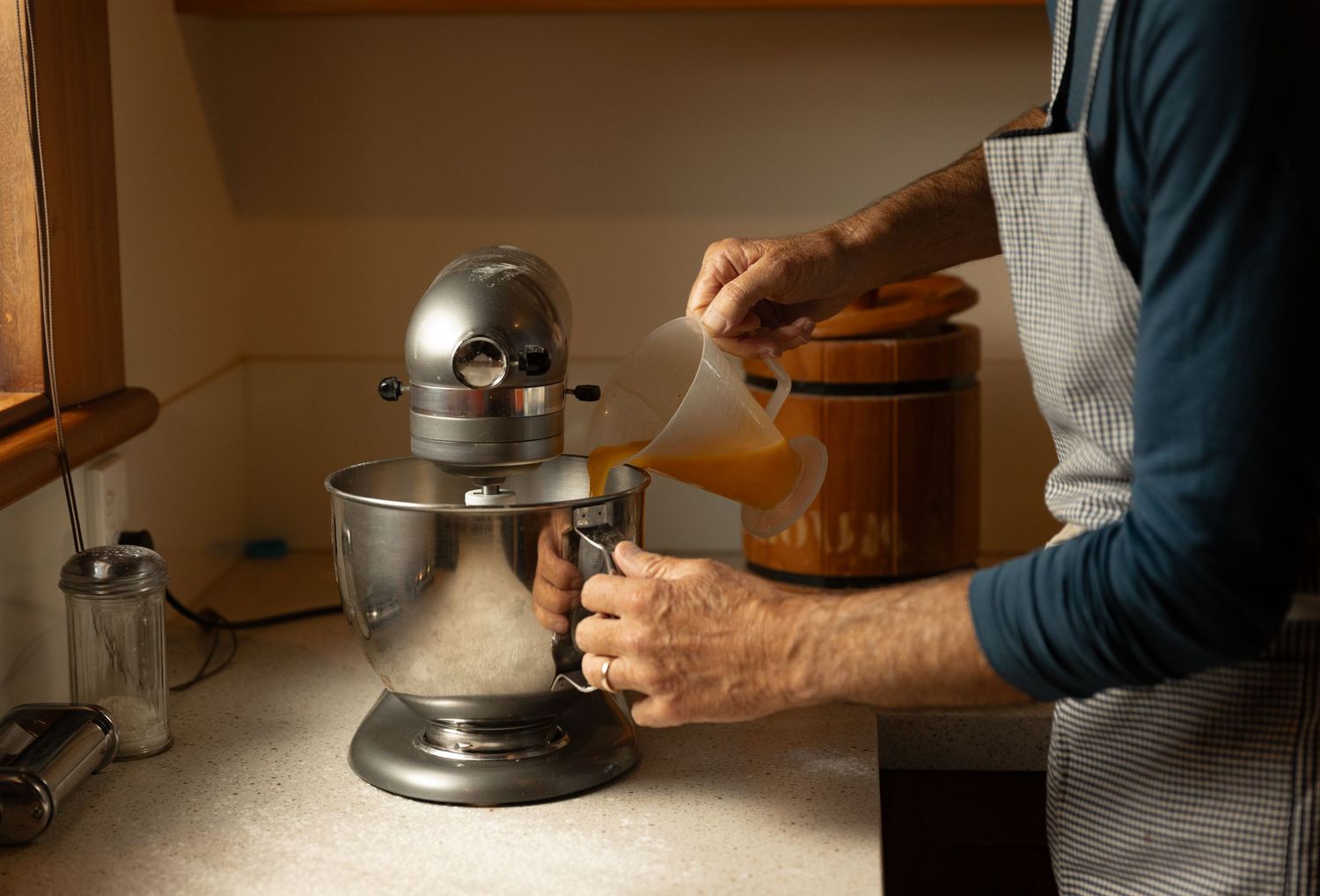 Person pouring liquid ingredient into a KitchenAid stand mixer bowl while baking in a kitchen.