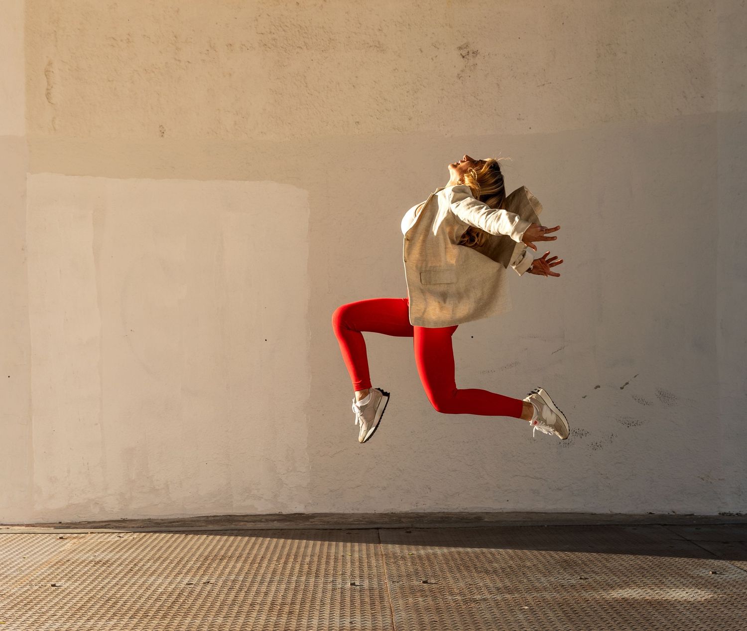 Woman in red leggings and white jacket jumping mid-air against a concrete wall.