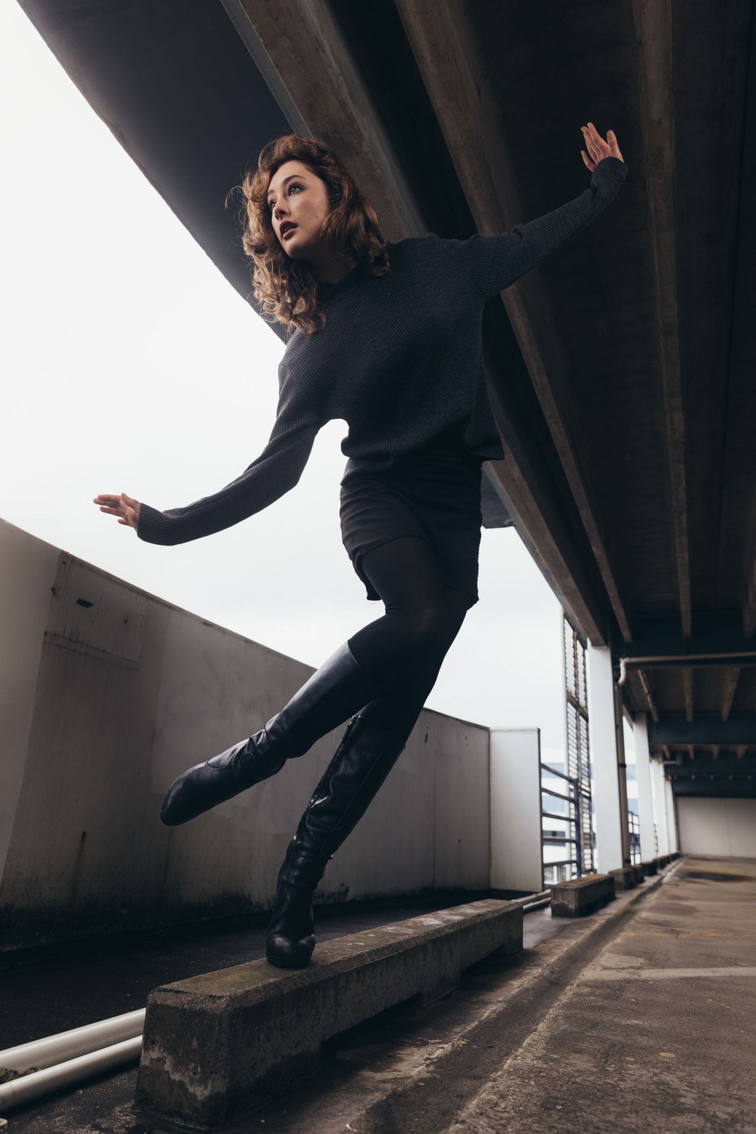 Female dancer in all-black attire striking a dynamic pose on concrete ledge under a bridge.
