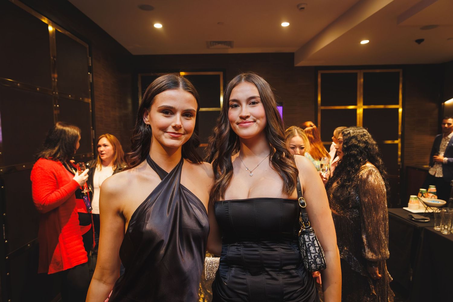 Two young women in black formal dresses smile together at an elegant evening awards event.