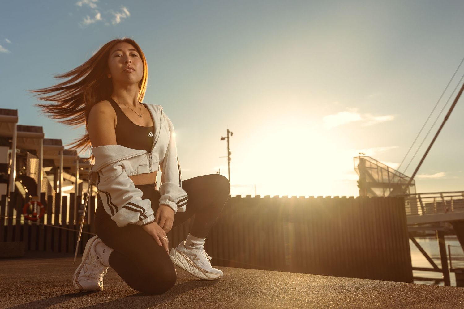 Woman in black sports bra and track pants kneeling on pavement with dramatic sunset backlight.