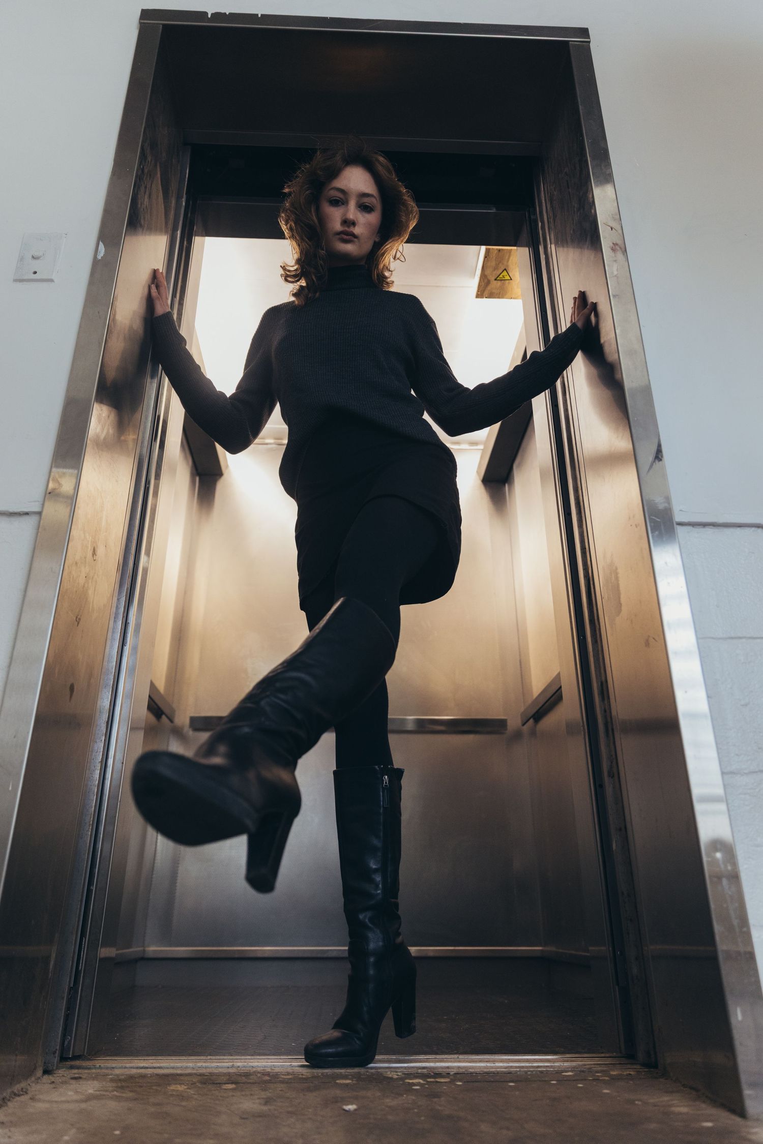 Woman in all-black outfit posing dramatically in open elevator doorway with one leg raised.
