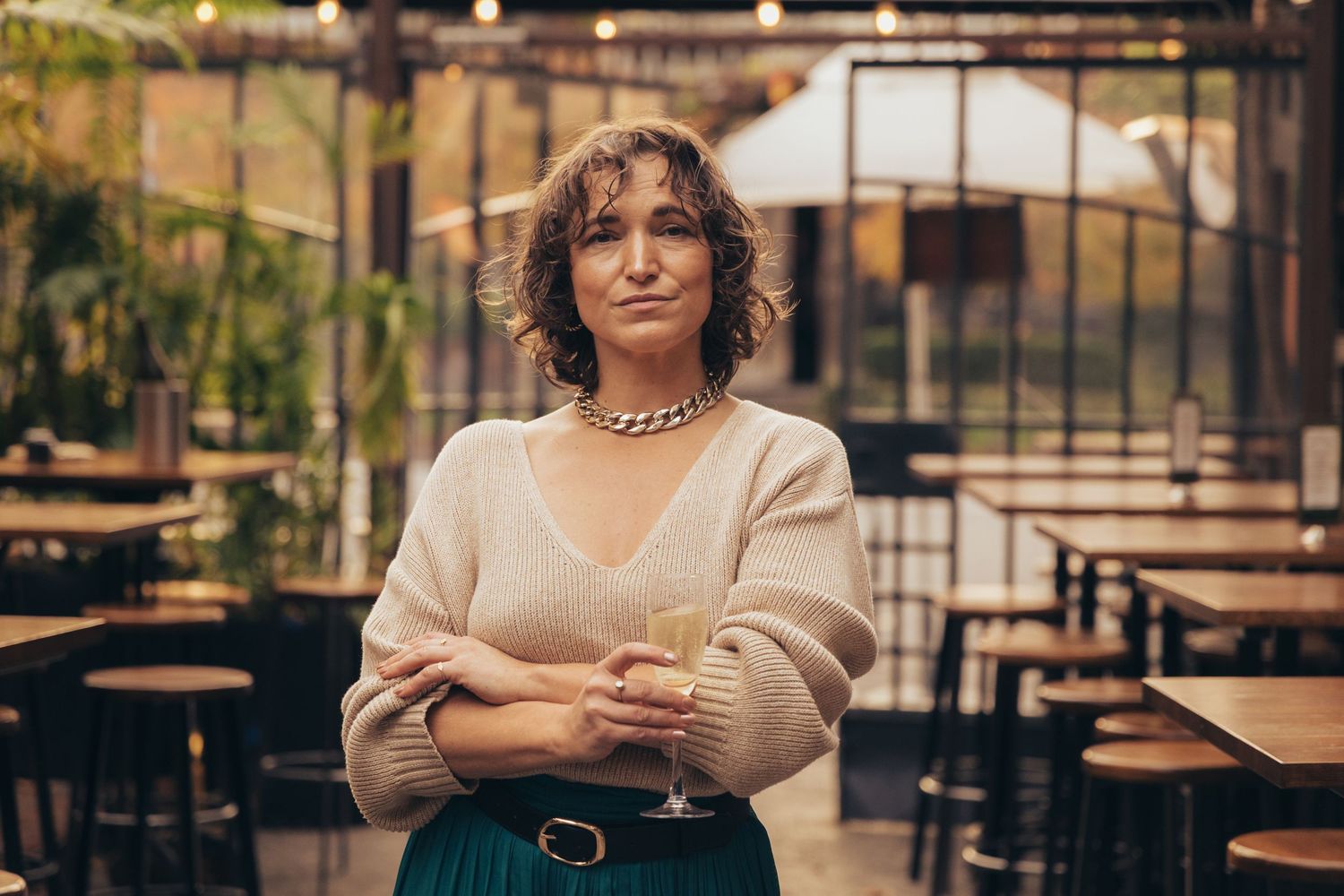 Woman holding wine glass with arms crossed in a stylish restaurant setting, wearing a beige sweater and green skirt.