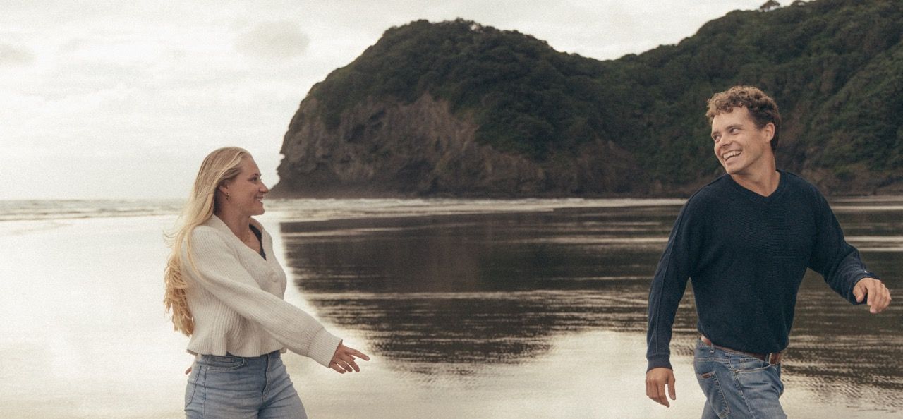 A couple walks hand-in-hand on a scenic beach with dramatic cliffs and calm waters in the background.