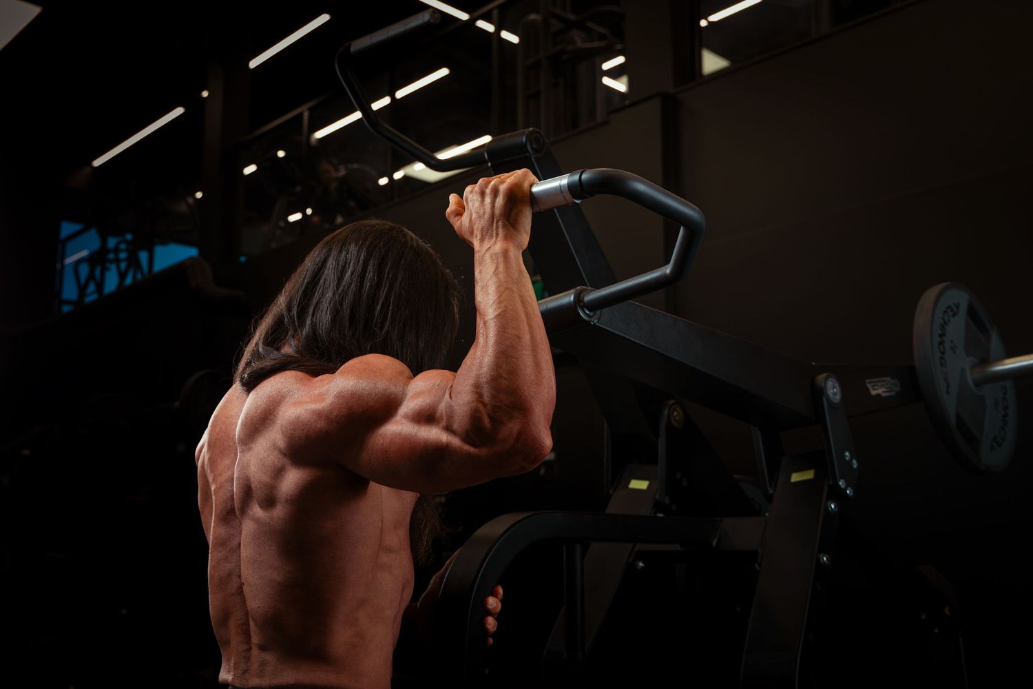 Muscular man performing pull-ups on gym bar, showing defined back muscles in dark fitness center.