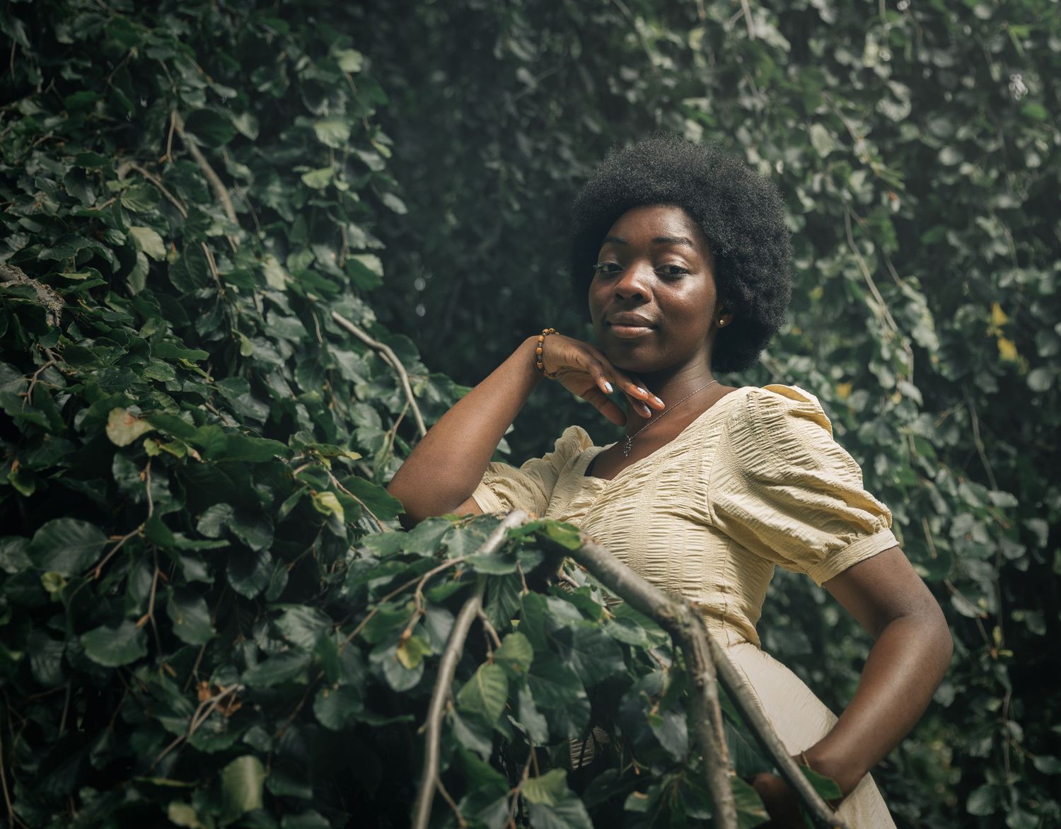 Young Black woman in yellow dress posing gracefully among lush green ivy leaves in natural light.