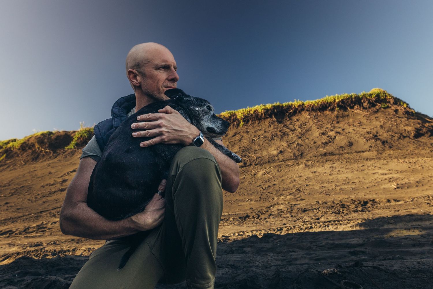 Bald man sitting outdoors holding small black dog against his chest on rocky hillside terrain.