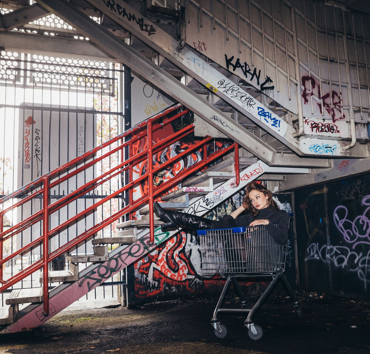 Young woman sits in shopping cart beneath graffiti-covered stairs in abandoned industrial building.