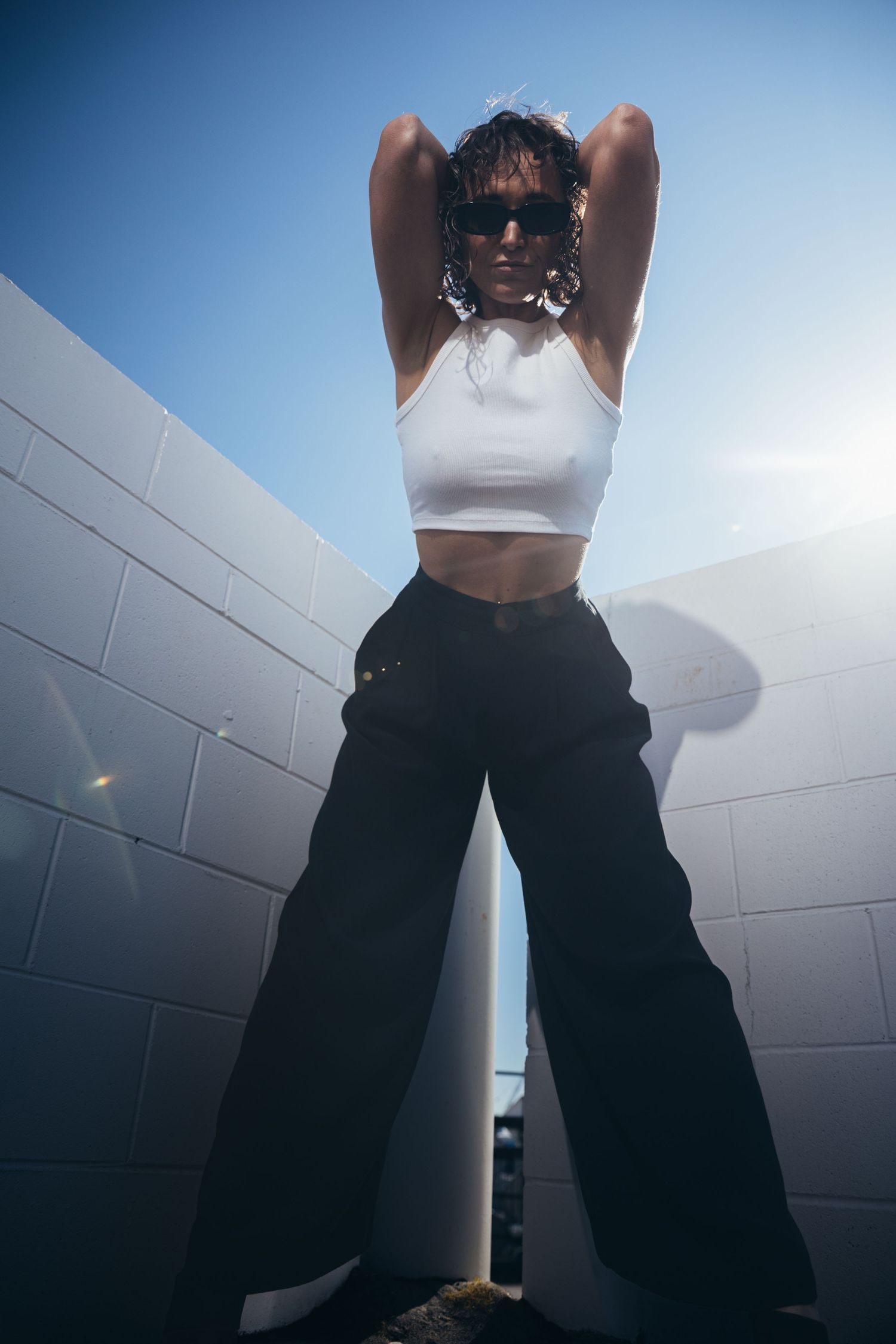 Woman in white crop top and black wide-leg pants poses with arms raised against white brick wall in sunlight.