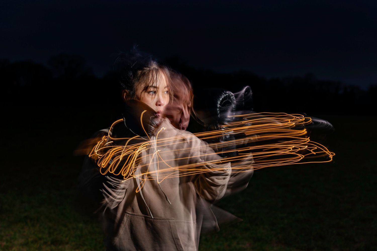 Woman in gray hoodie surrounded by swirling orange light painting trails in darkness.