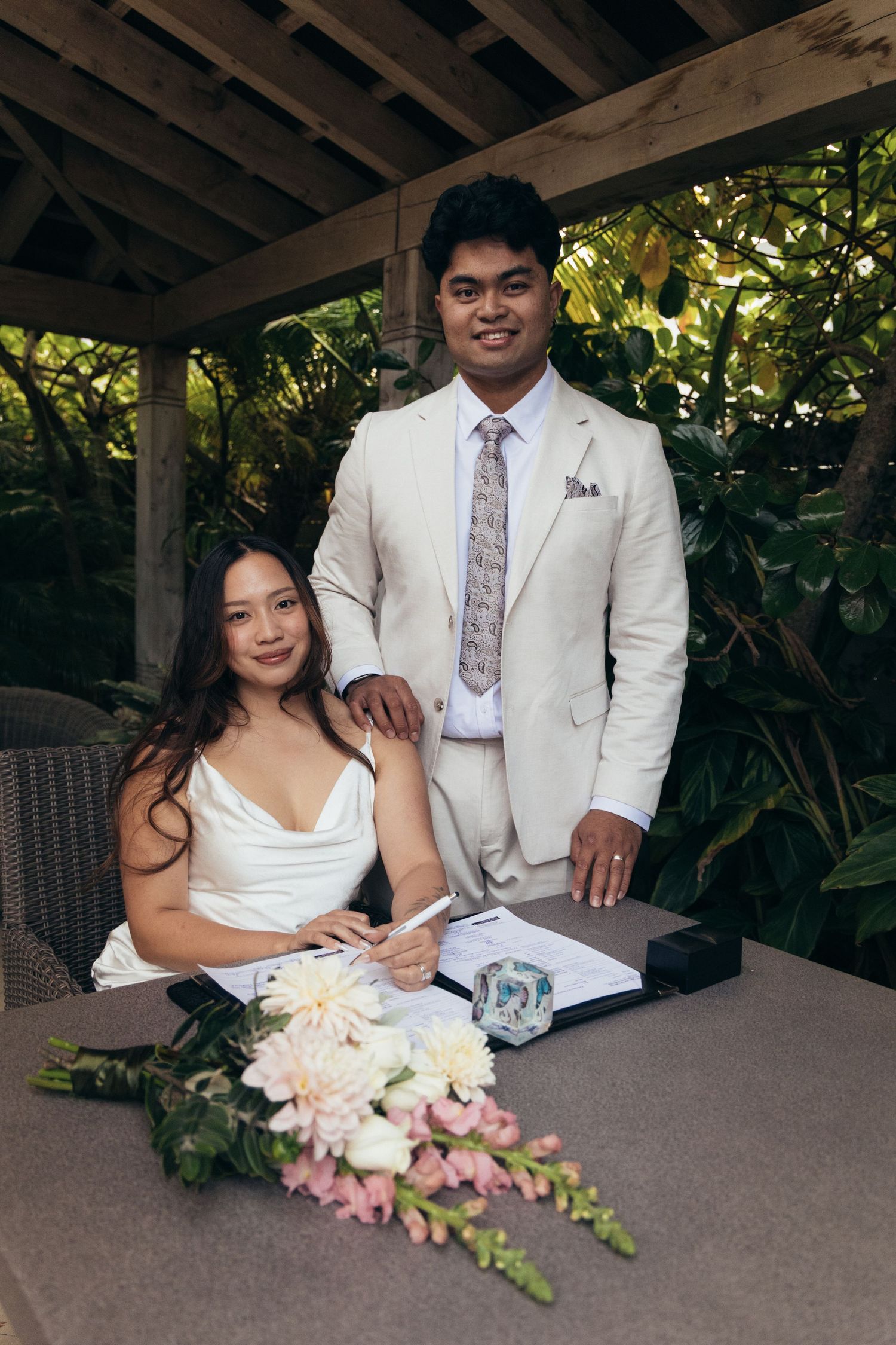Bride signs wedding documents while groom stands beside her at a tropical outdoor ceremony venue.
