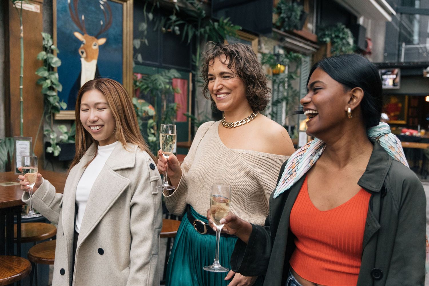 Three diverse women smiling and holding champagne glasses in a decorated pub interior.