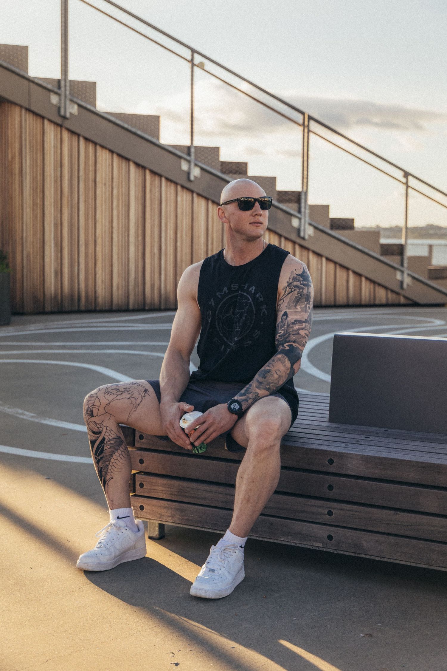 Tattooed bald man in black tank top and shorts sitting on bench at outdoor basketball court wearing sunglasses.