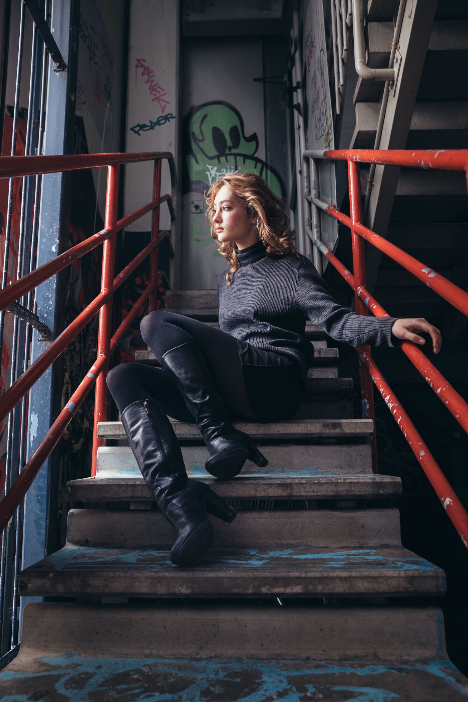 Young woman in gray sweater and black boots sits on graffiti-covered staircase with red railings.