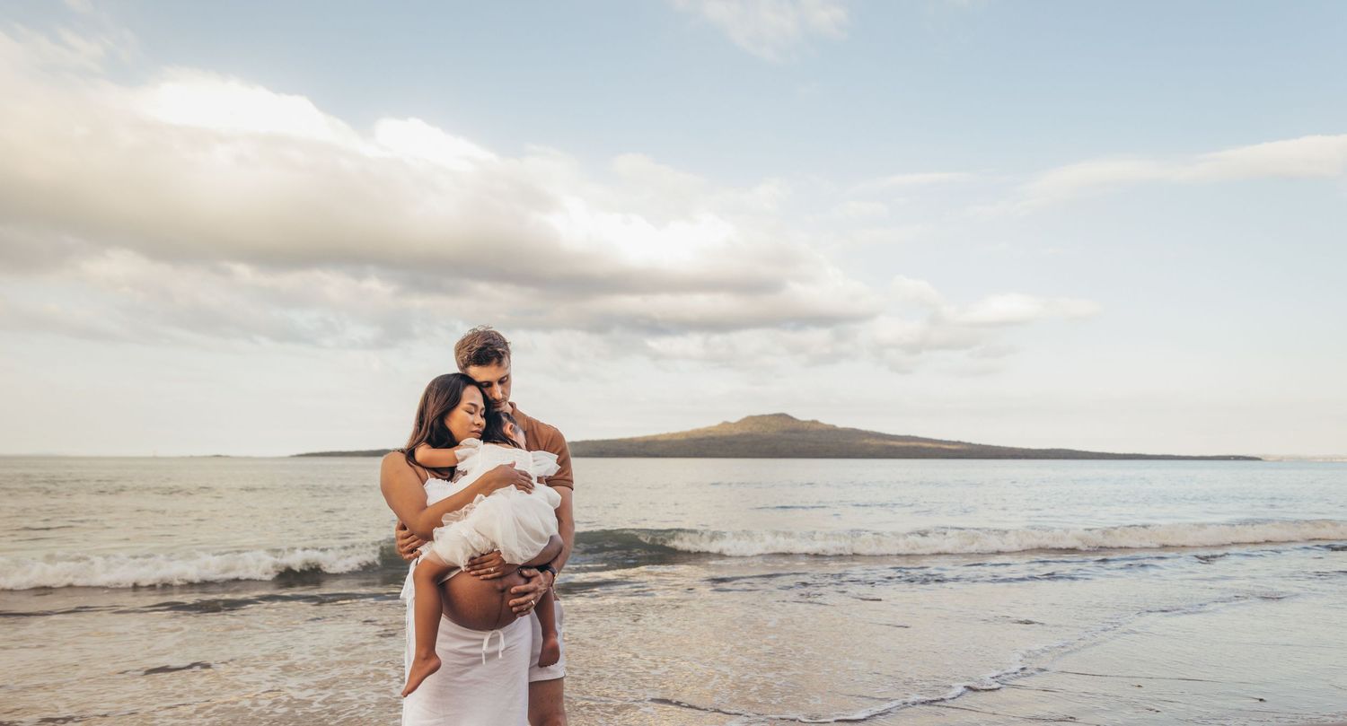 A couple embraces on a serene beach with a volcanic island in the background under a cloudy sky.