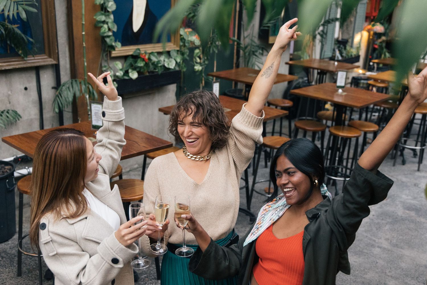Three diverse women celebrating with champagne glasses raised in a trendy restaurant bar setting.