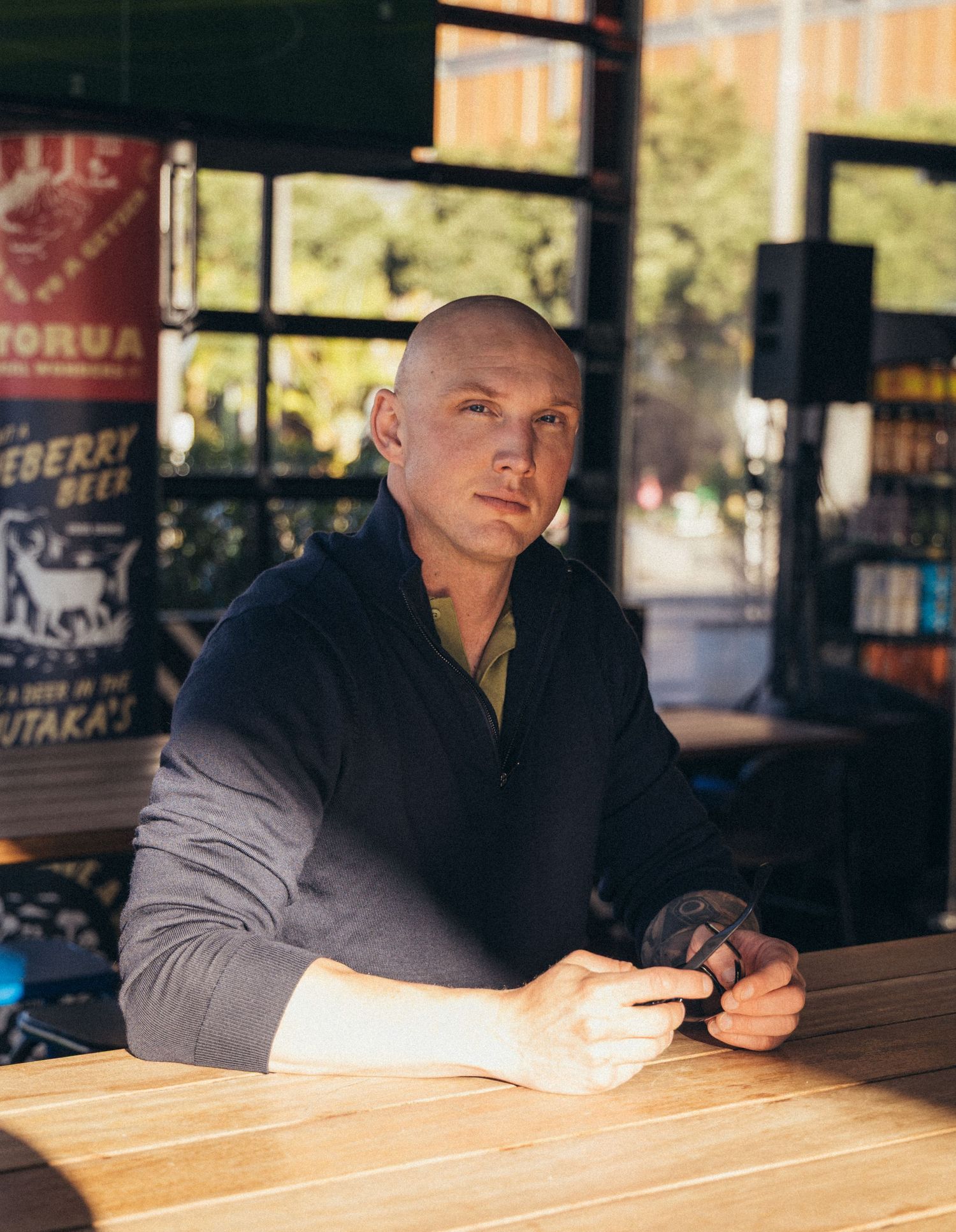 Bald man in dark sweater sitting at wooden bar table holding drink in casual setting.