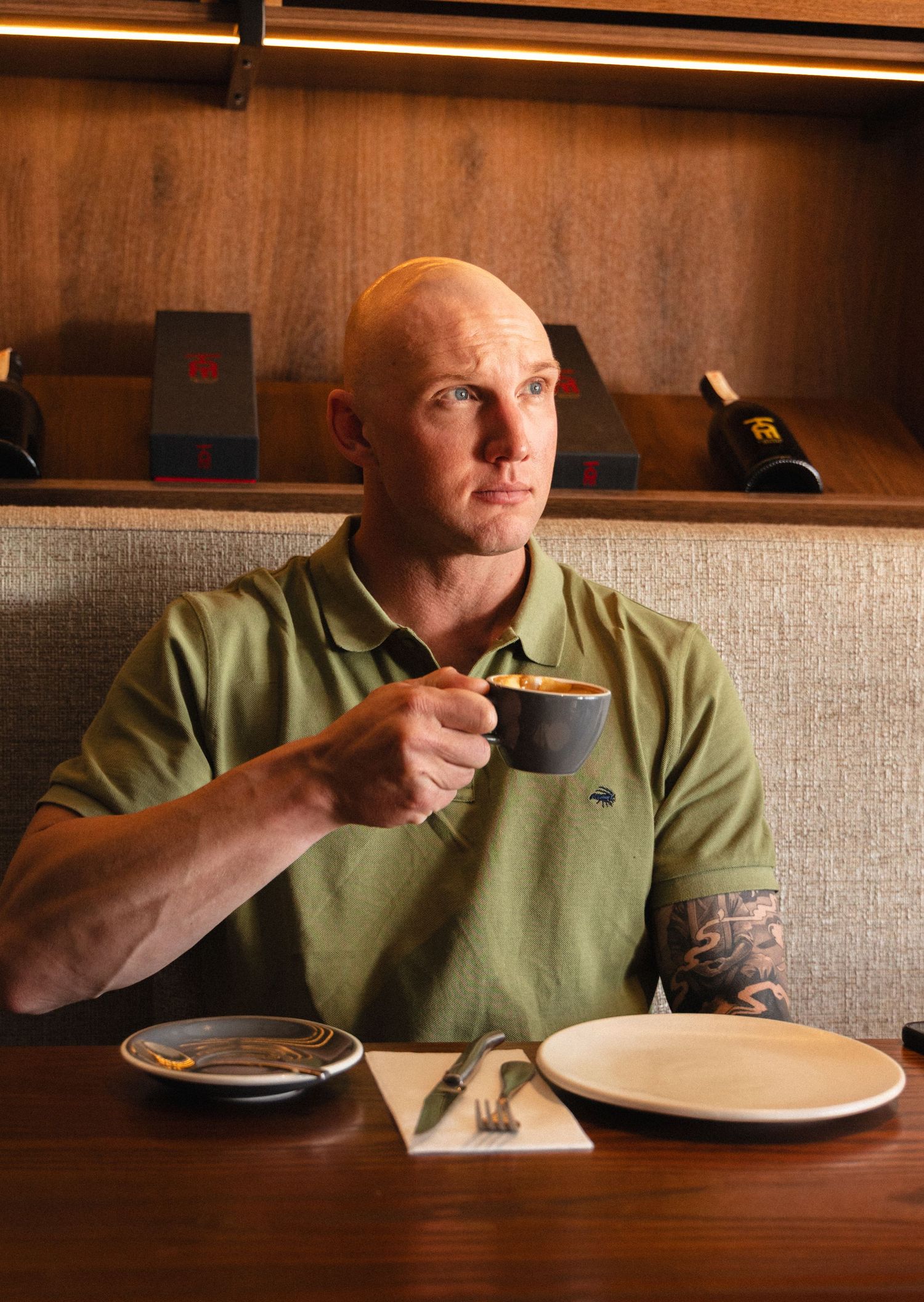 Bald man with tattoo sleeve sipping espresso at restaurant booth with wine bottle and plates.
