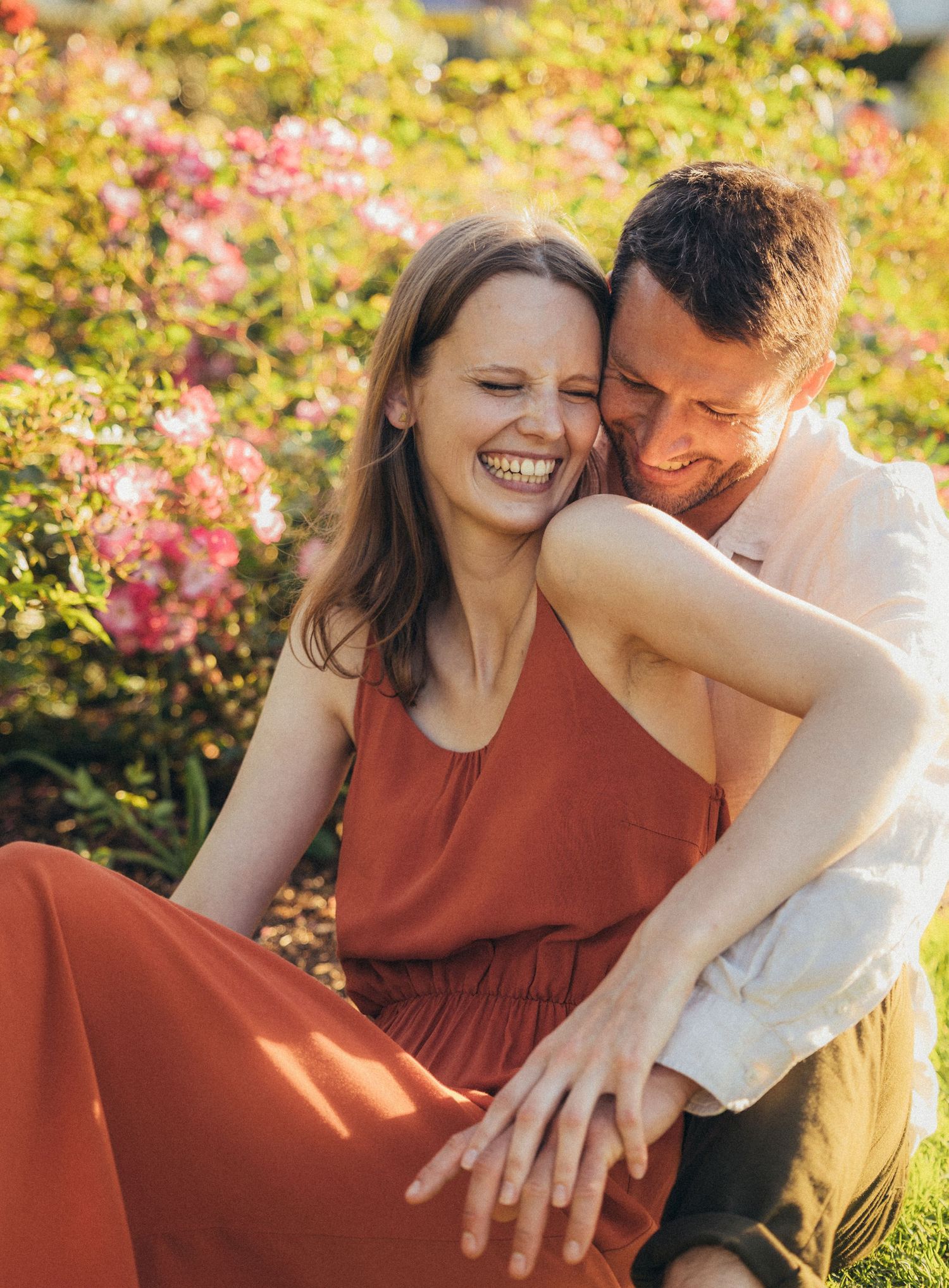 Joyful couple embracing in a sunlit garden with blooming pink flowers, woman in rust dress laughing warmly.