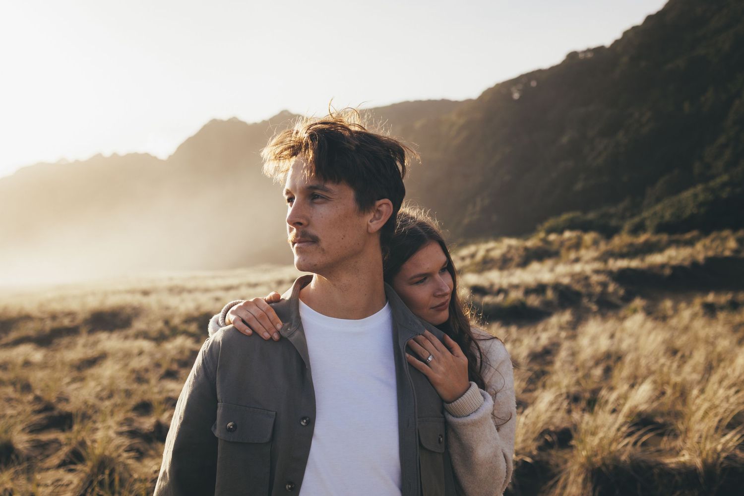 Young woman resting her head on a mans shoulder in a sunlit coastal field.