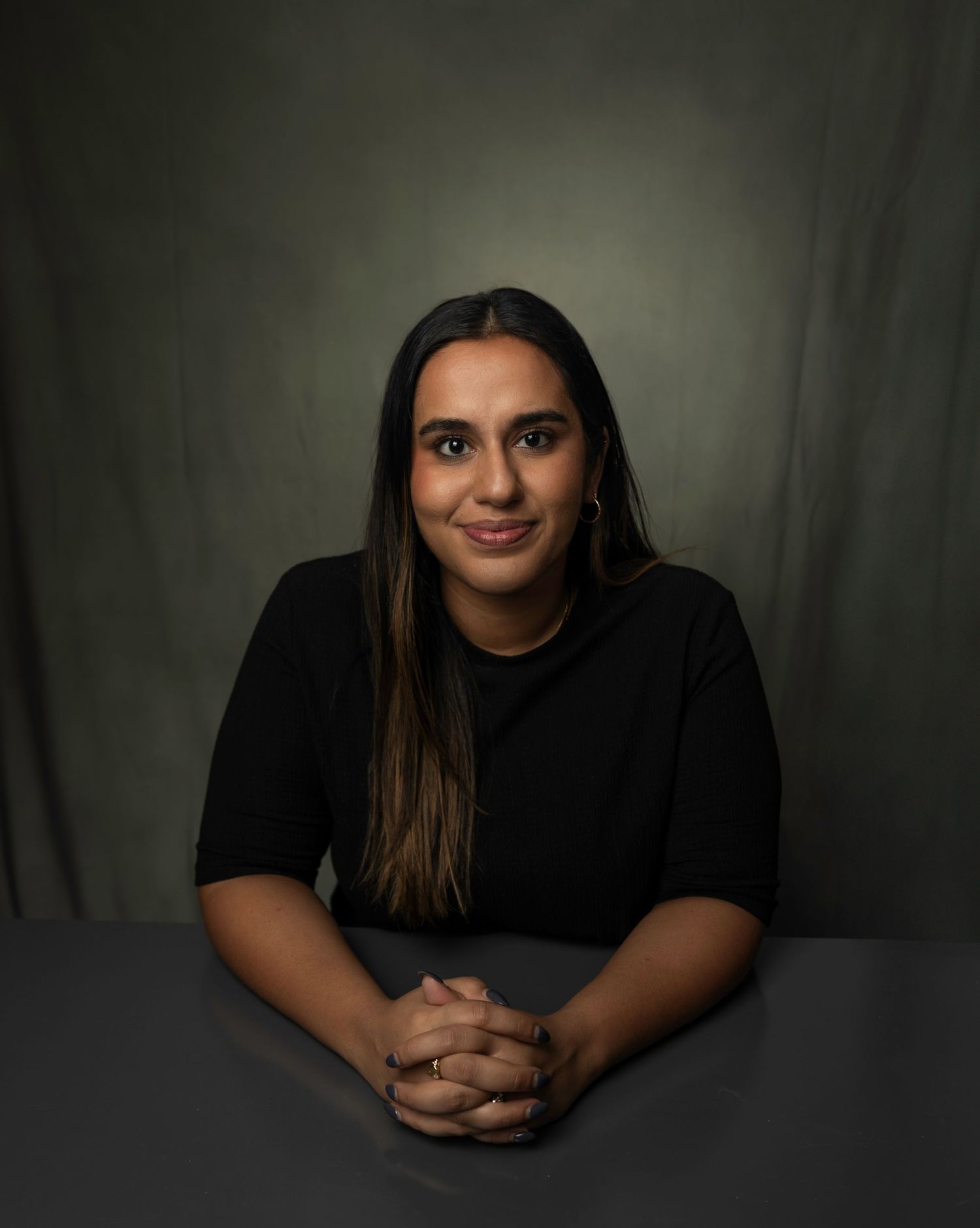 Woman in black top sits at table with hands clasped, smiling against dark green backdrop.