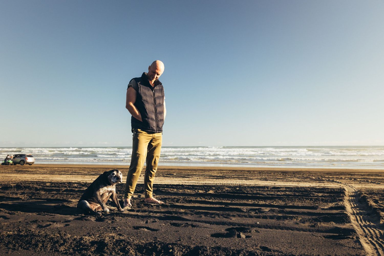 Bald man in puffer vest standing on beach with his small black dog on a clear day.