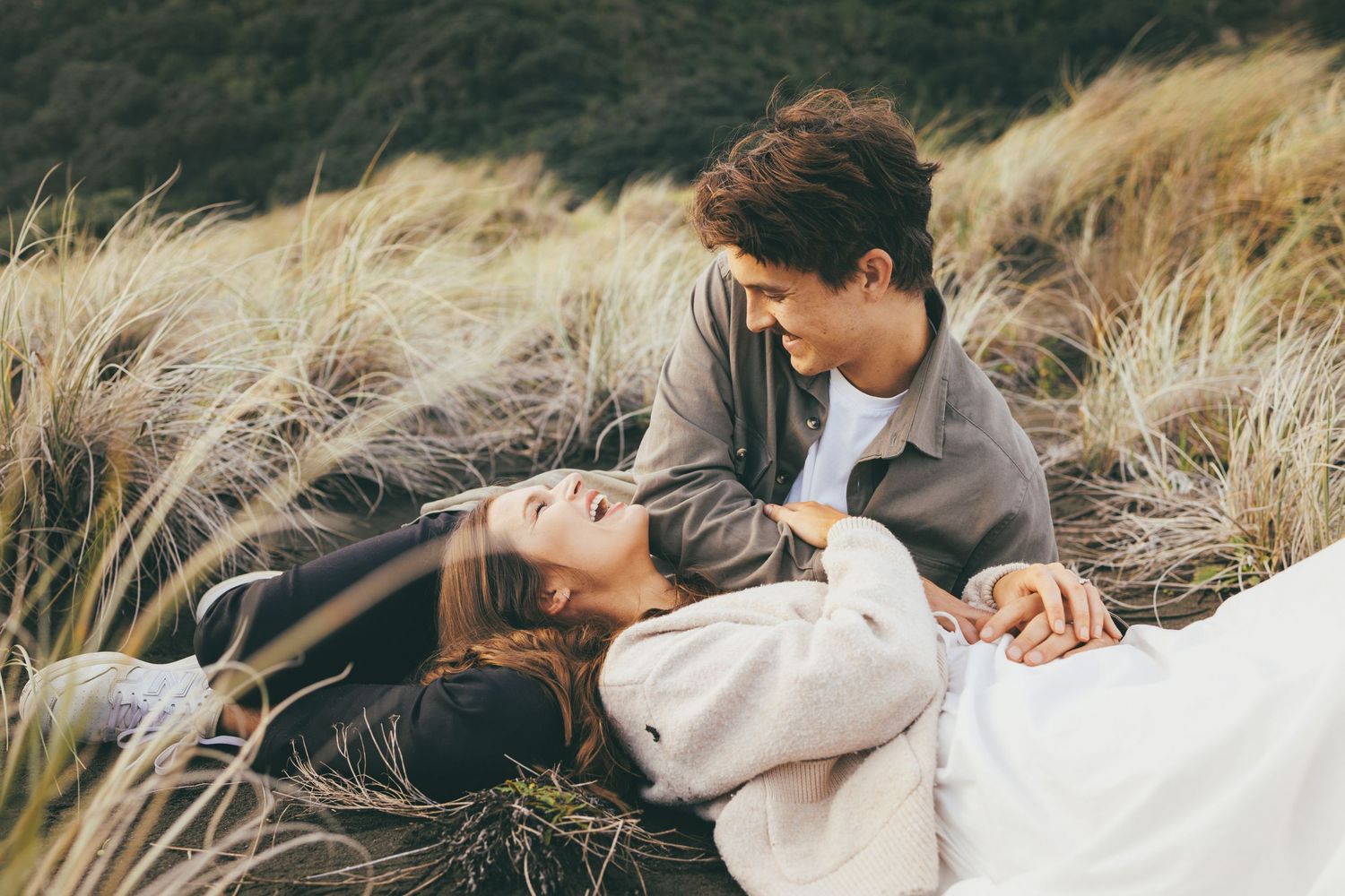 A young couple sharing a romantic moment lying in tall golden grass, smiling and gazing at each other outdoors.