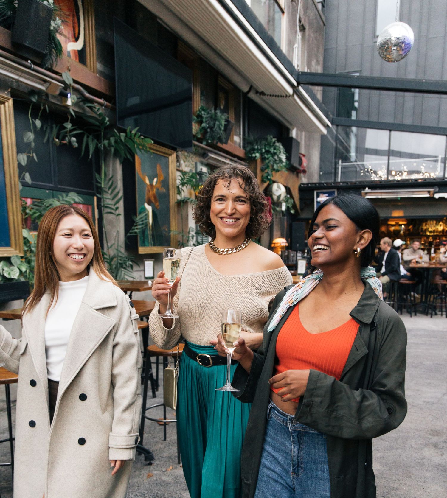Three smiling women holding champagne glasses outside a decorated restaurant entrance on a city street.