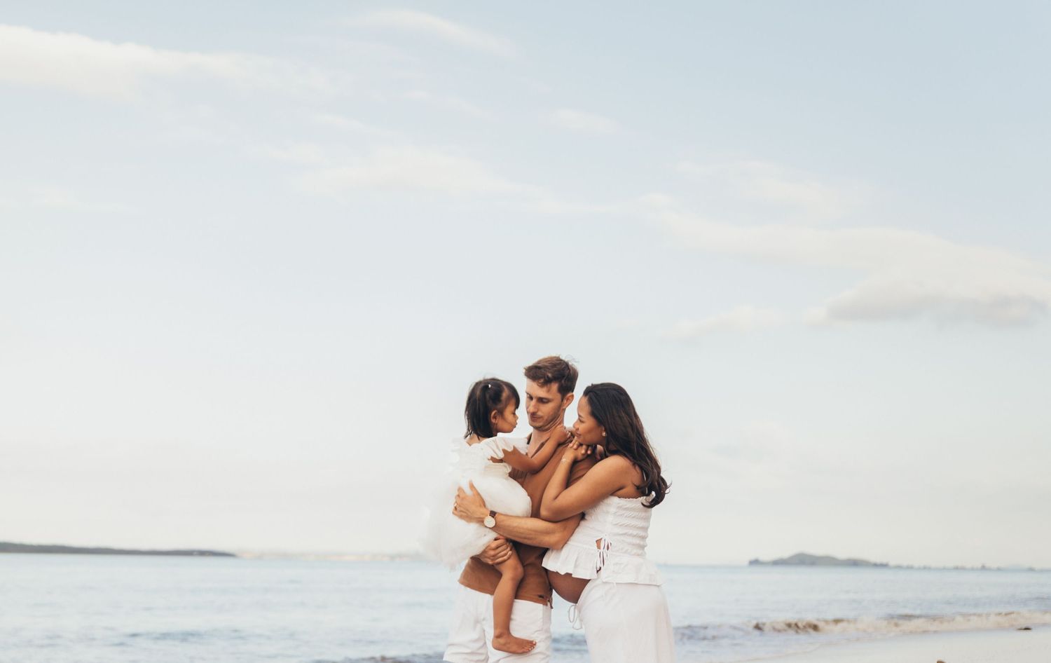 A happy family of three enjoys a sunny beach day, with parents holding their young child near the ocean shoreline.