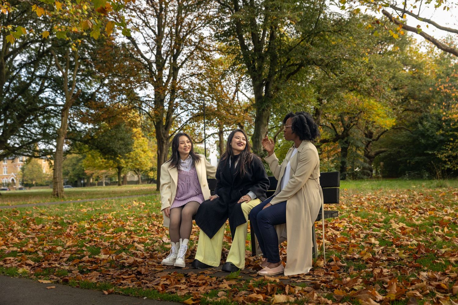 Three women sitting on a bench laughing together in a park surrounded by autumn foliage.
