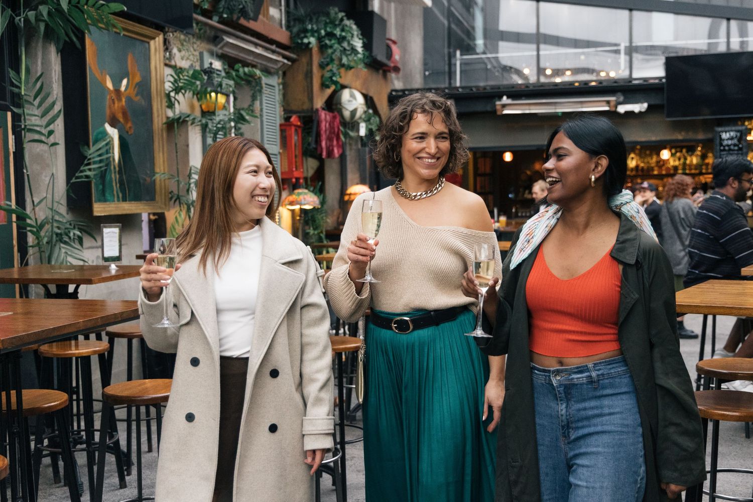 Three stylish women walking and holding champagne glasses in a decorated indoor market bar.